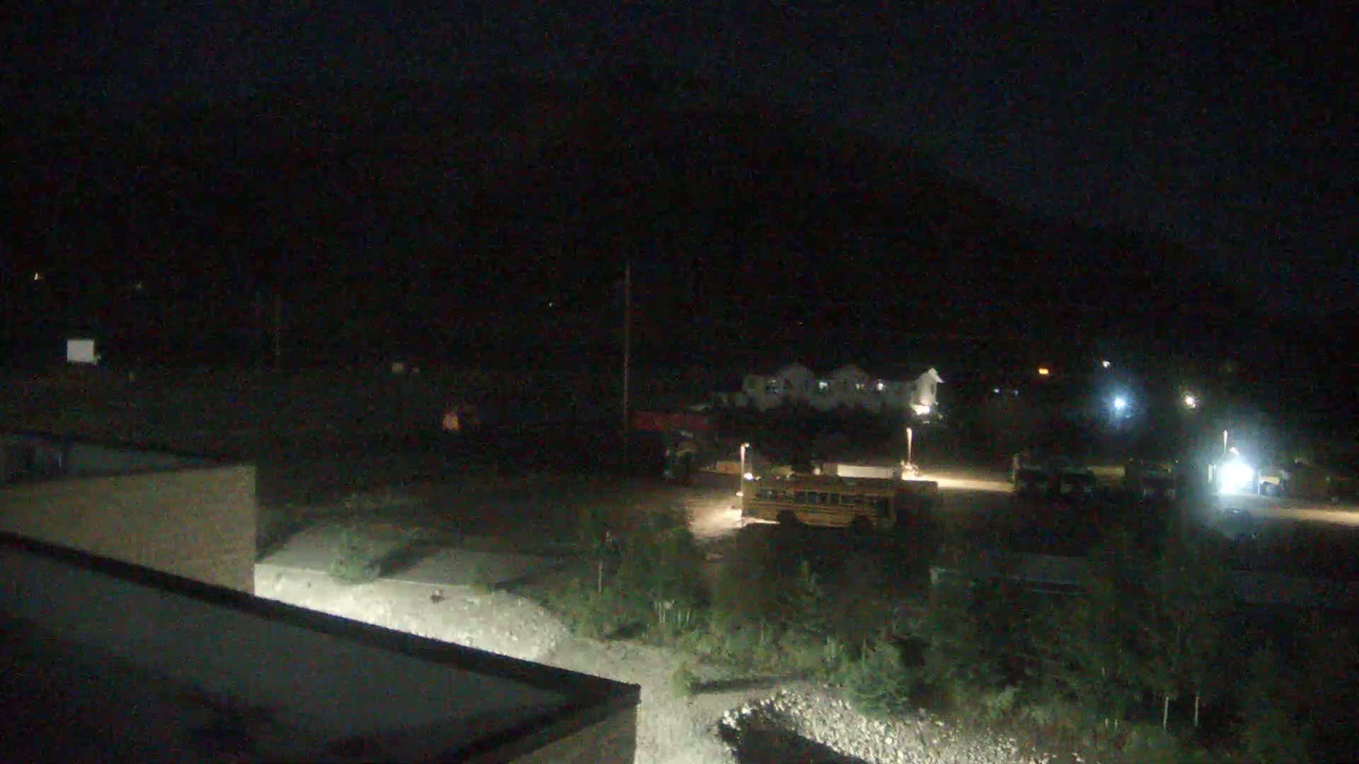 A nighttime, low-light view shows a school bus parked in a mostly empty lot with a few other vehicles, near some buildings and sparse vegetation, under a dark sky.