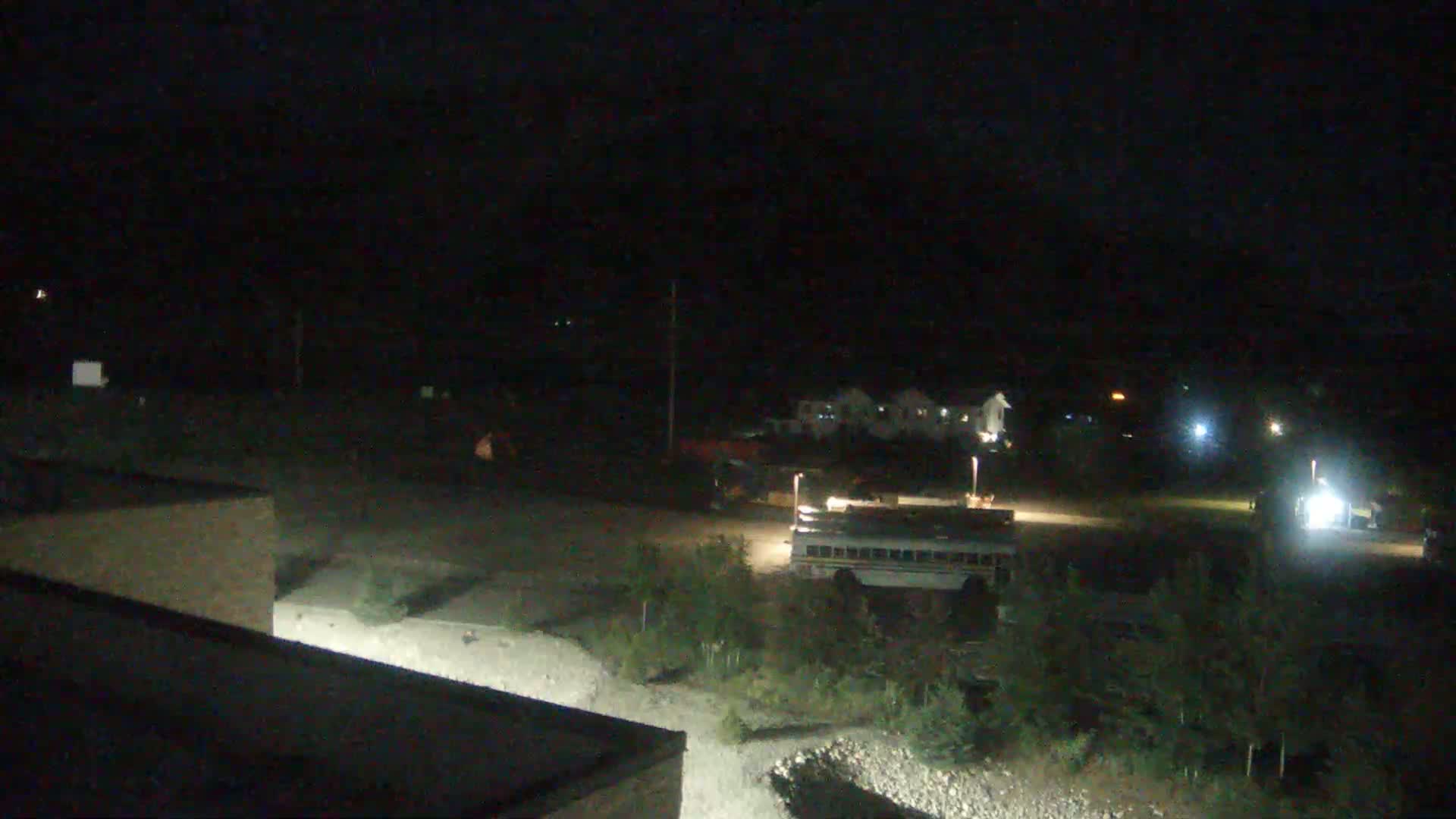 A nighttime, outdoor scene shows a school bus parked in a gravel lot near some buildings and low vegetation under a dark sky.