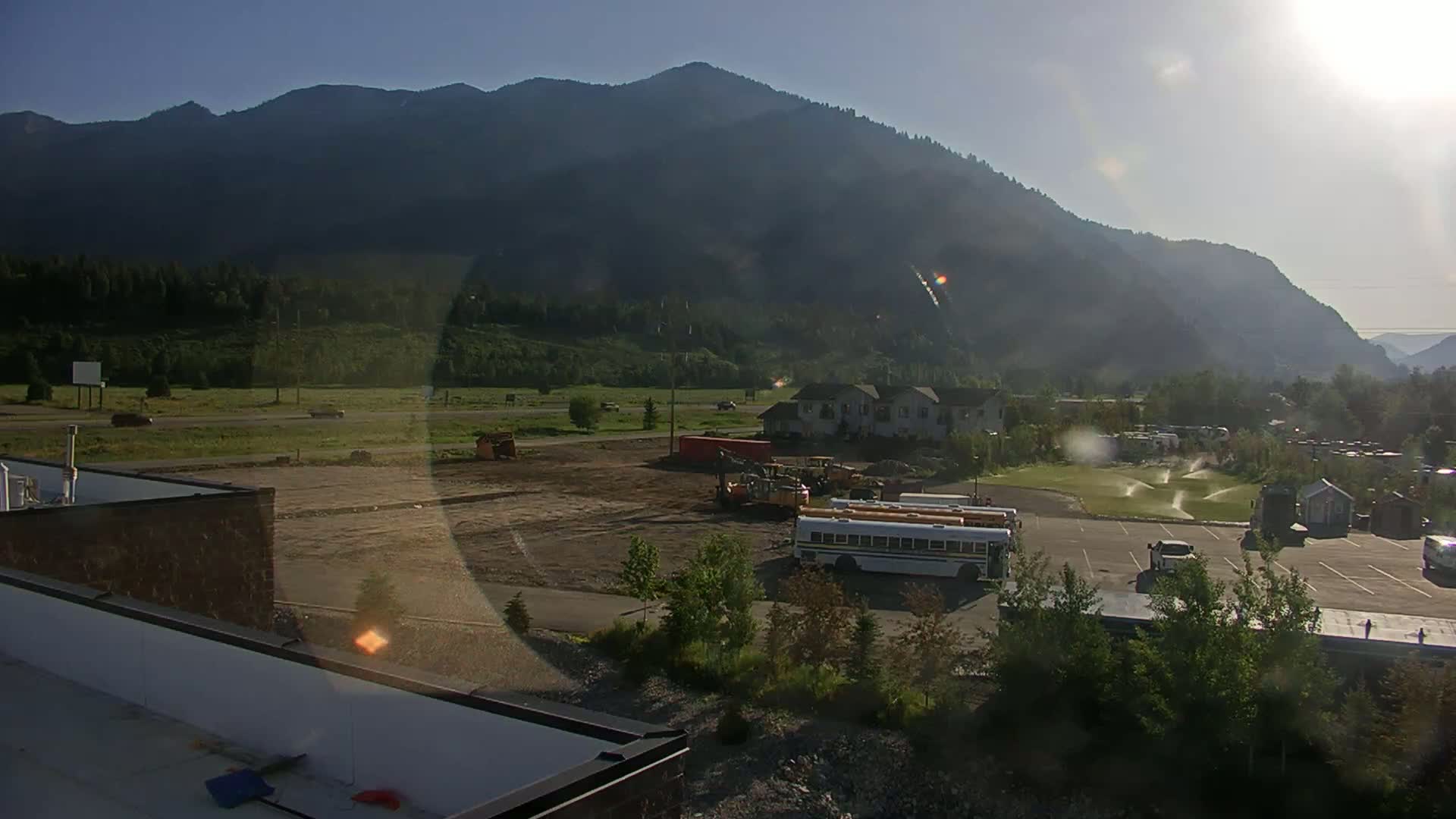 A sunny day reveals a construction site with earth-moving equipment near a parking lot containing a school bus, all set against a backdrop of mountains and lush green vegetation.