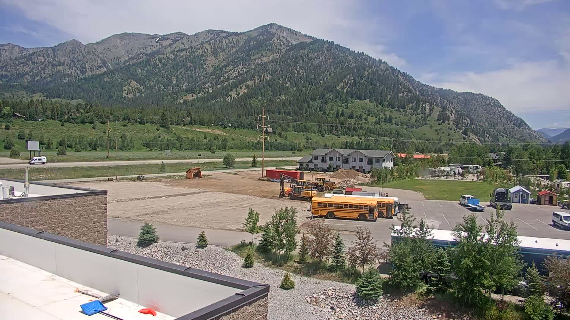 A sunny day reveals a construction site with earth-moving equipment near a parking lot containing a school bus, all set against a backdrop of mountains and lush green vegetation.