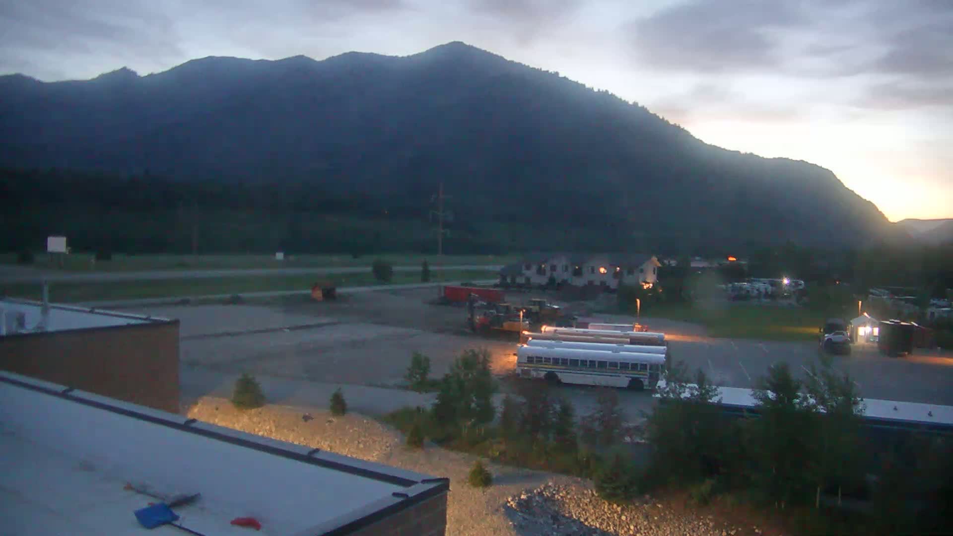 A dusk-lit parking lot with school buses and construction equipment is situated in front of a large building and a mountain range under an overcast sky.