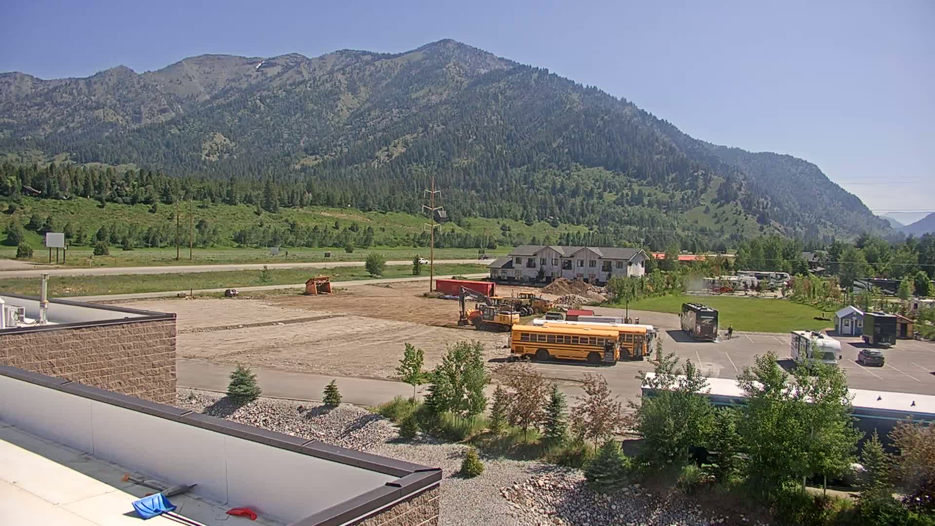 A construction site with heavy machinery and school buses sits in a valley below a large mountain range on a sunny day.