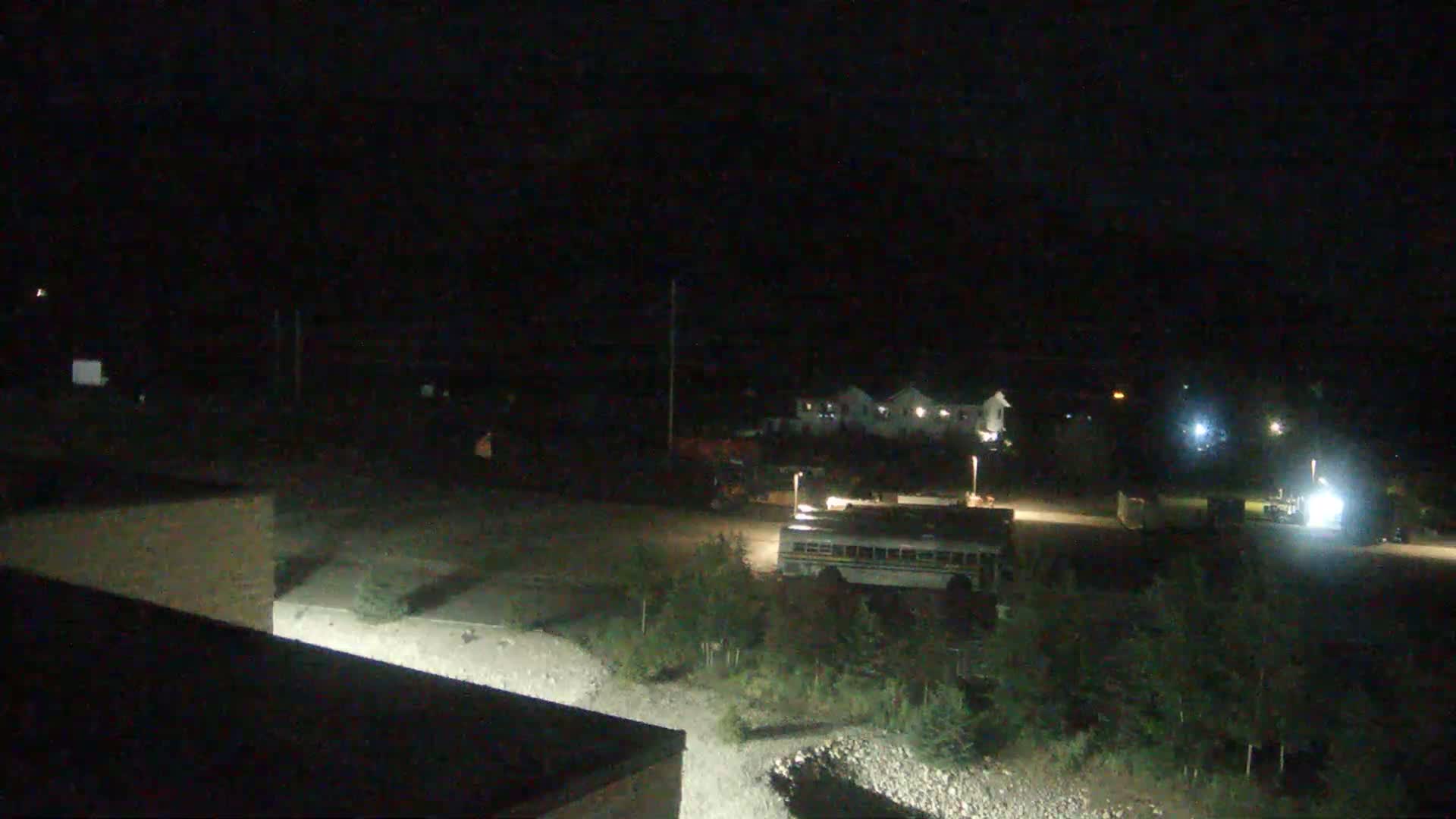 A nighttime, low-light view of a mostly empty parking lot with a school bus, some vehicles, and a few buildings in the background.