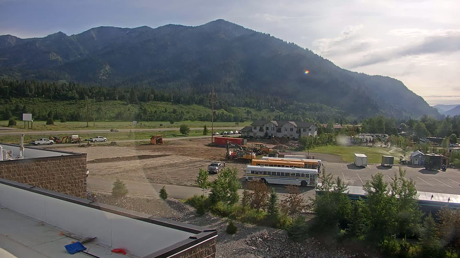 A construction site with heavy machinery and a school bus is situated in a valley next to a large mountain under a partly cloudy sky.