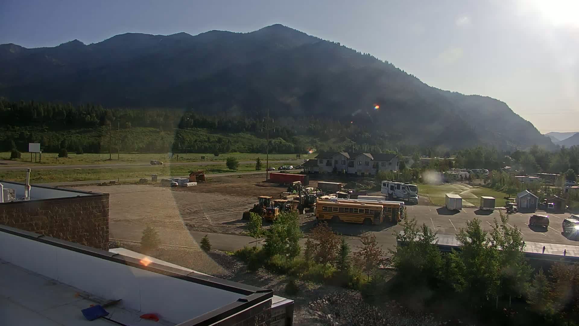 A sunny construction site with heavy machinery, a school bus, and residential buildings is situated in a valley before a large mountain range.