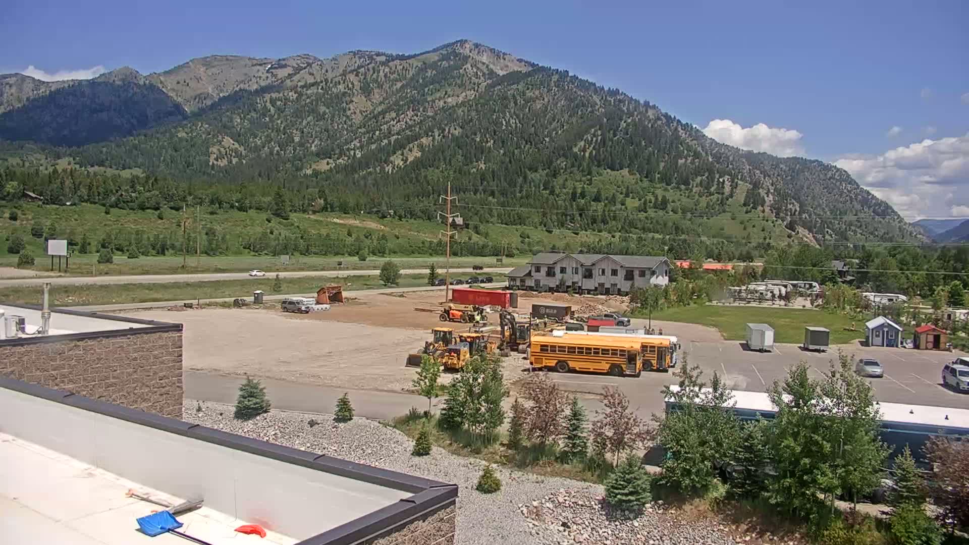 A construction site with heavy machinery and a school bus is situated in a valley, overlooked by a mountain range under a mostly sunny sky.