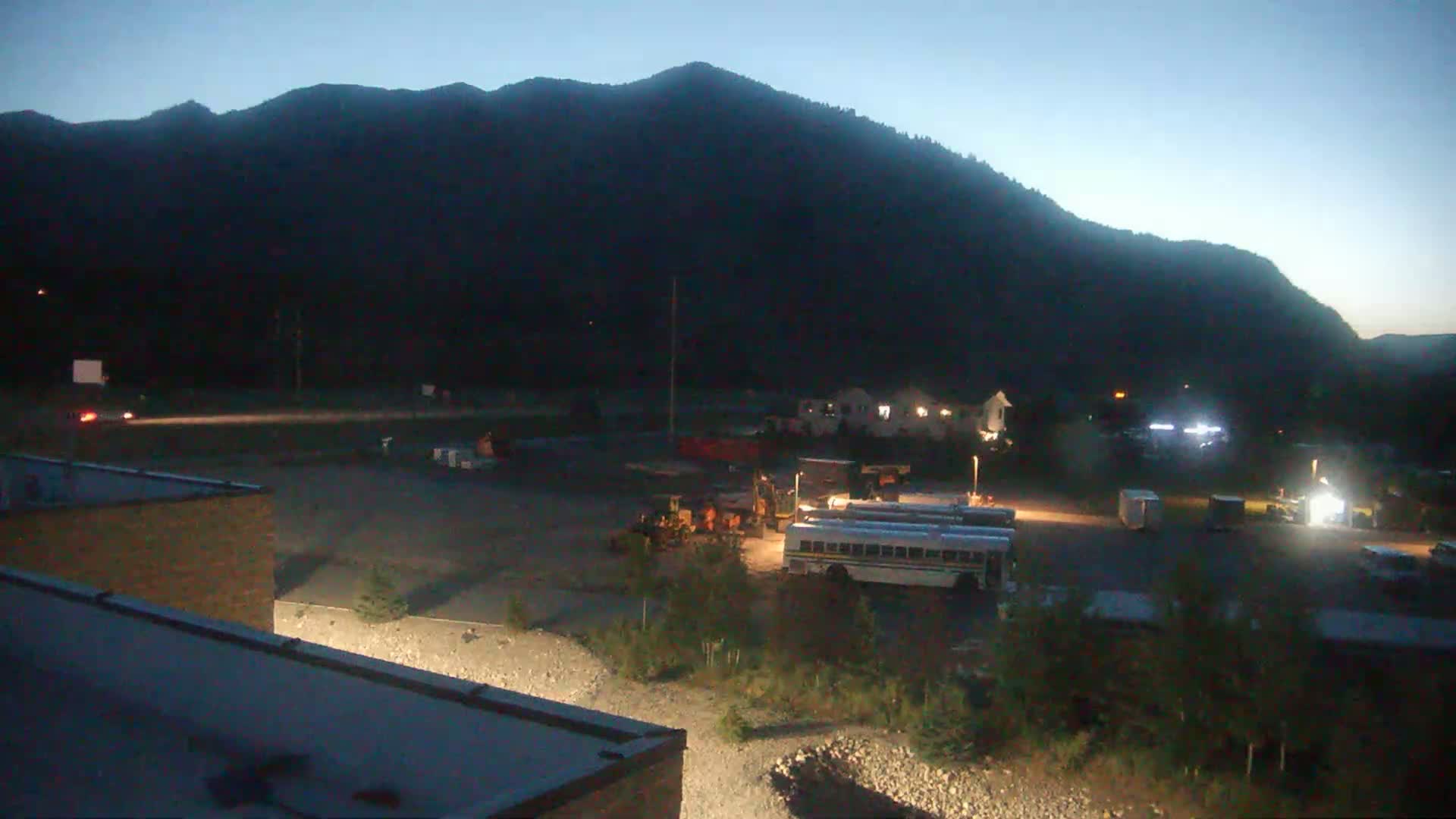 Under a twilight sky, construction or maintenance equipment and several buses are parked in a lot near a dark mountain range.