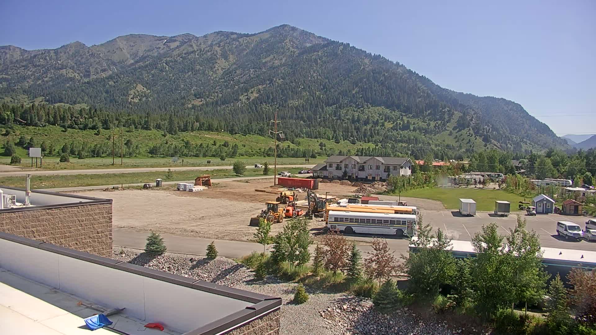 Construction equipment sits on a cleared lot near a two-story building, with a school bus parked nearby, all under a clear, sunny sky in front of a mountain range.