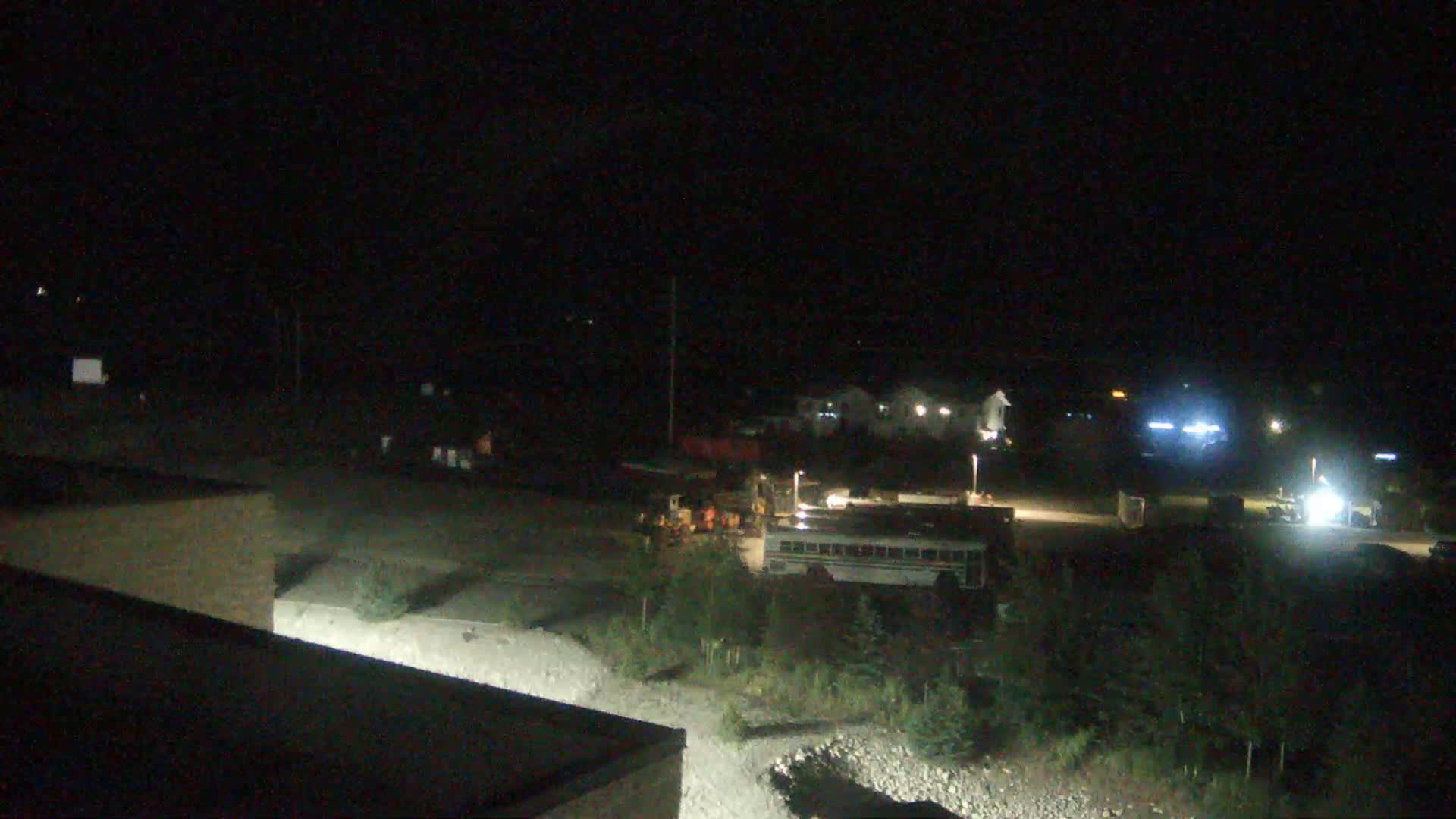 A nighttime, dimly lit scene shows a school bus parked near construction equipment and buildings in a sparsely vegetated area.