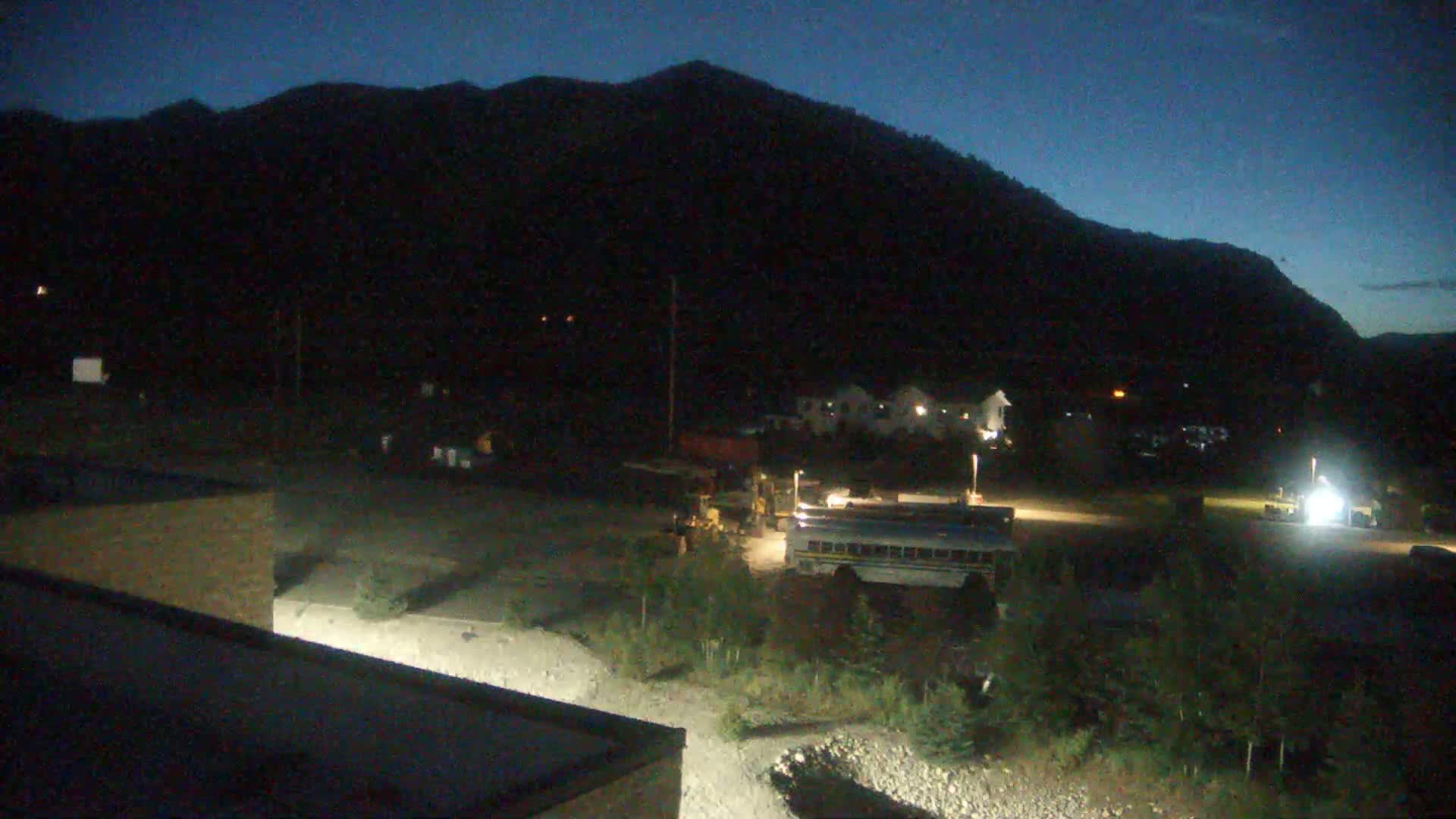 At night, under a dark blue sky, heavy machinery and a school bus are parked in a lot near a mountain, with low-lying vegetation in the foreground.