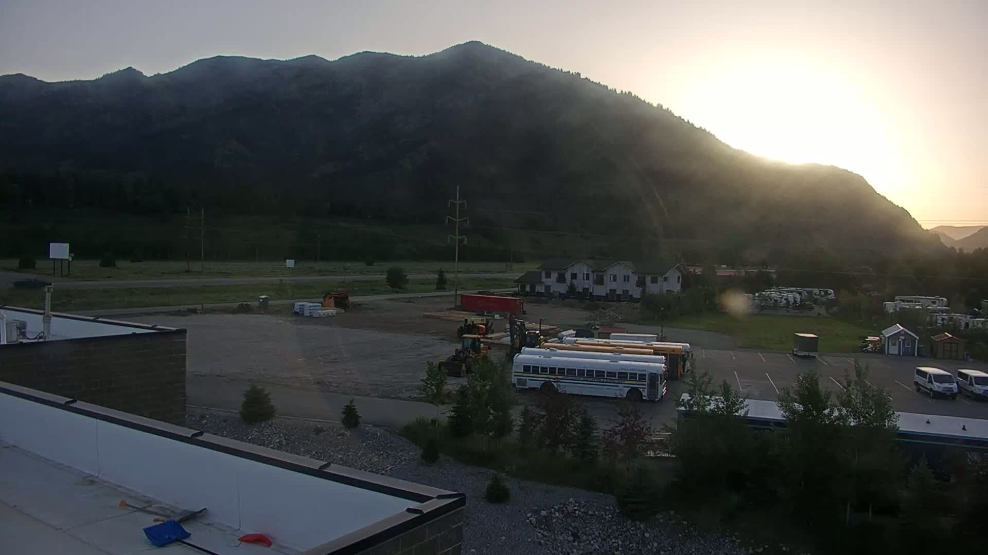 A sunrise illuminates a mountainside behind a flat, paved area containing construction equipment, several school buses, and some small buildings, with a few trees and shrubs scattered throughout.