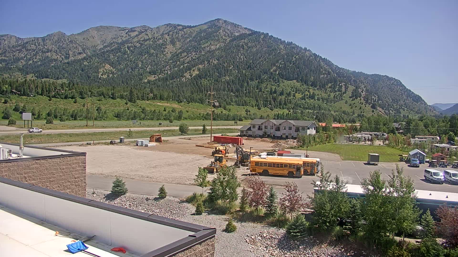 Construction equipment is operating on a cleared lot near a highway and a mountain range under a clear, sunny sky.