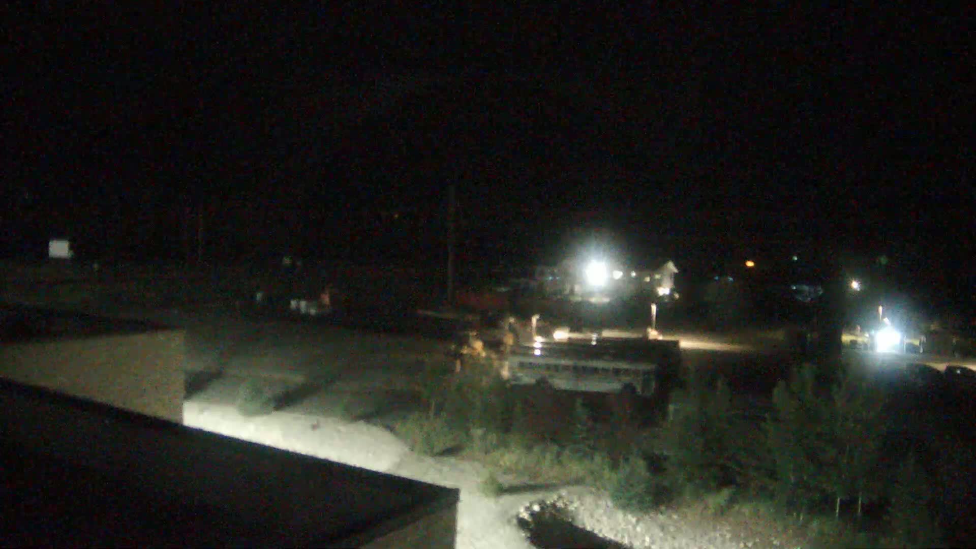 A nighttime, outdoor scene shows a parking lot with a bus and other vehicles, some lights, and sparse vegetation.