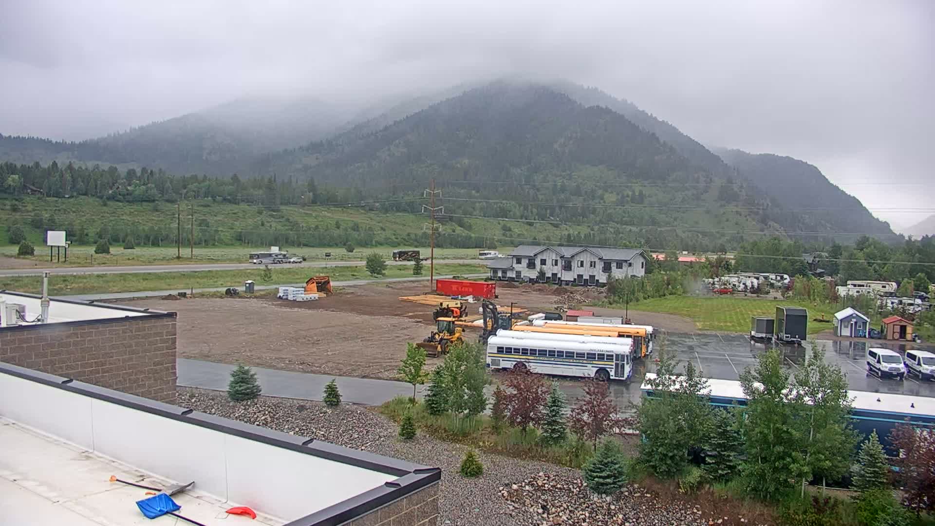 A construction site with heavy machinery and school buses is situated near a residential building and a highway, all set against a backdrop of misty mountains.