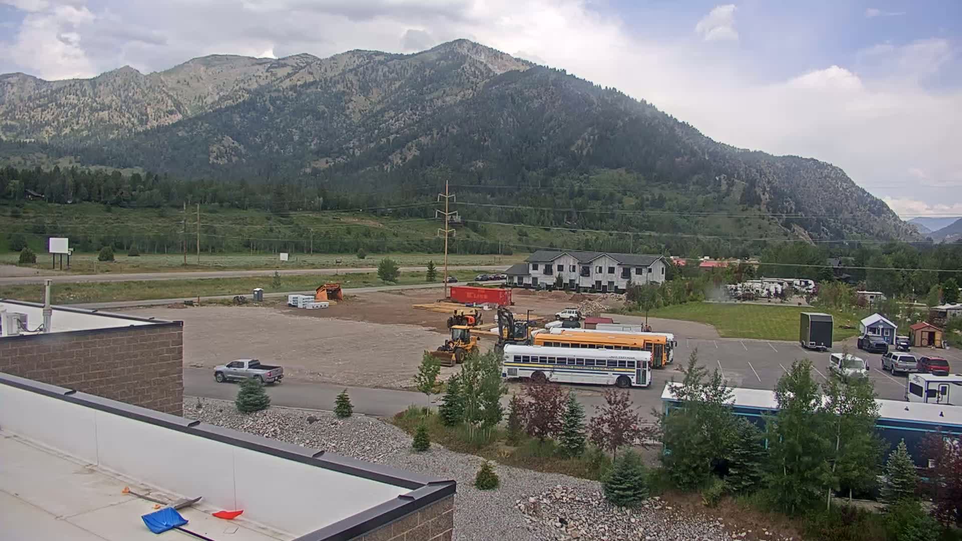A construction site with heavy machinery and a school bus is visible in a parking lot at the base of a mountain range under partly cloudy skies.