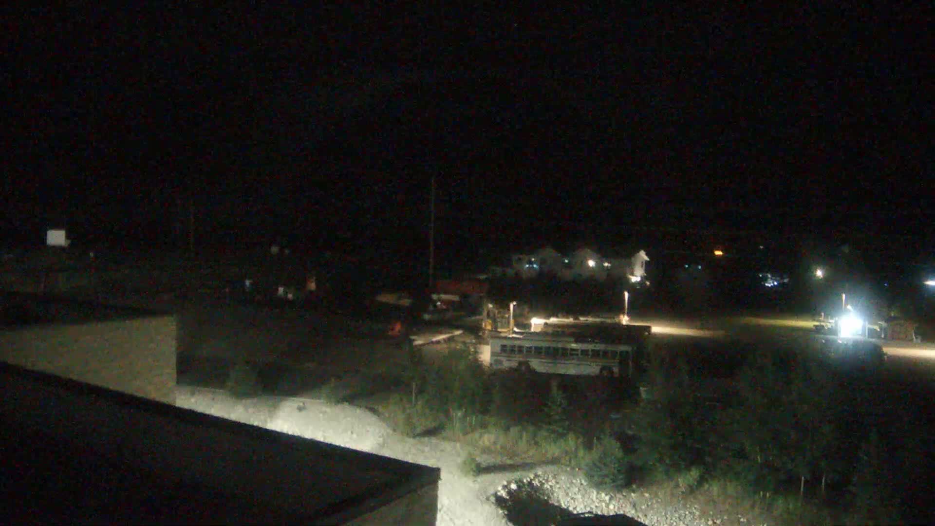 A nighttime, low-light view shows a school bus and other vehicles parked in a gravel lot near some buildings and vegetation.