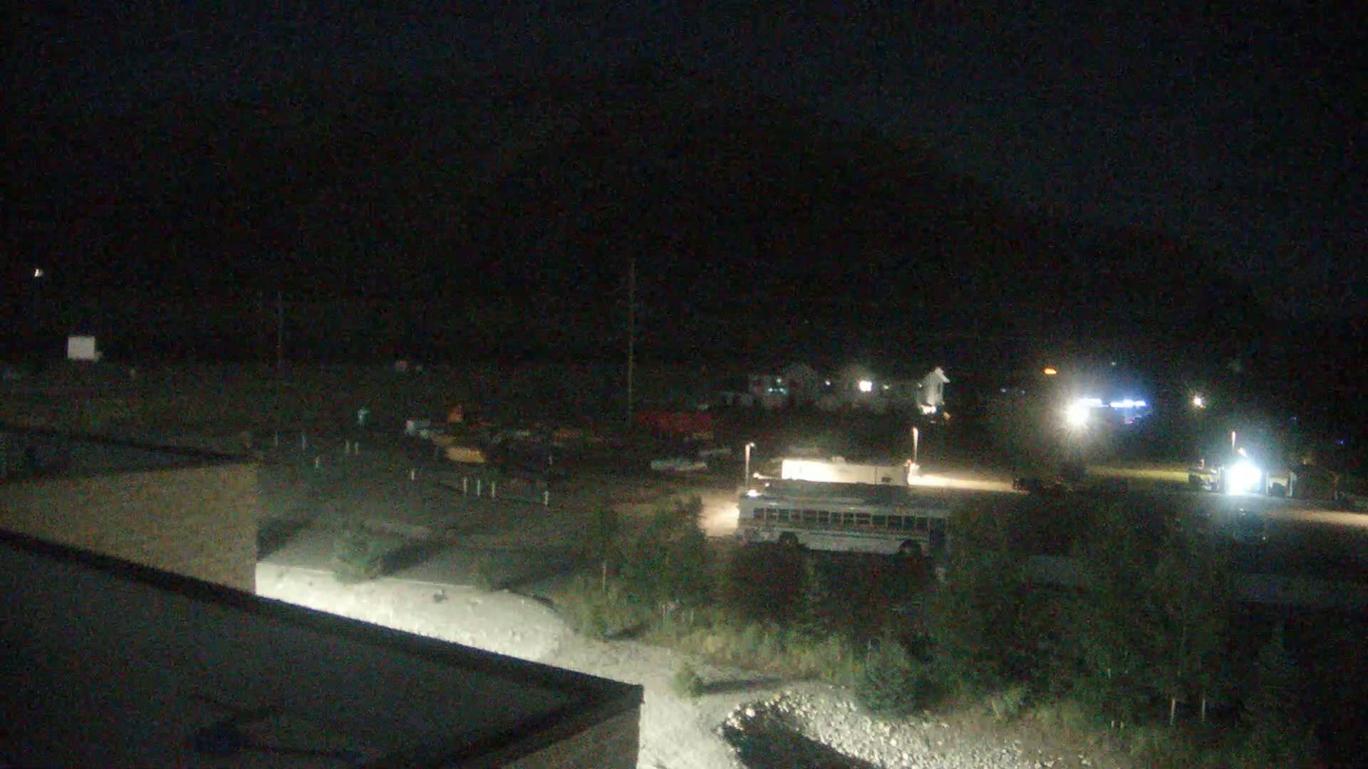 A nighttime, outdoor view shows a school bus parked in a lot with other vehicles, some buildings in the distance under a dark sky.