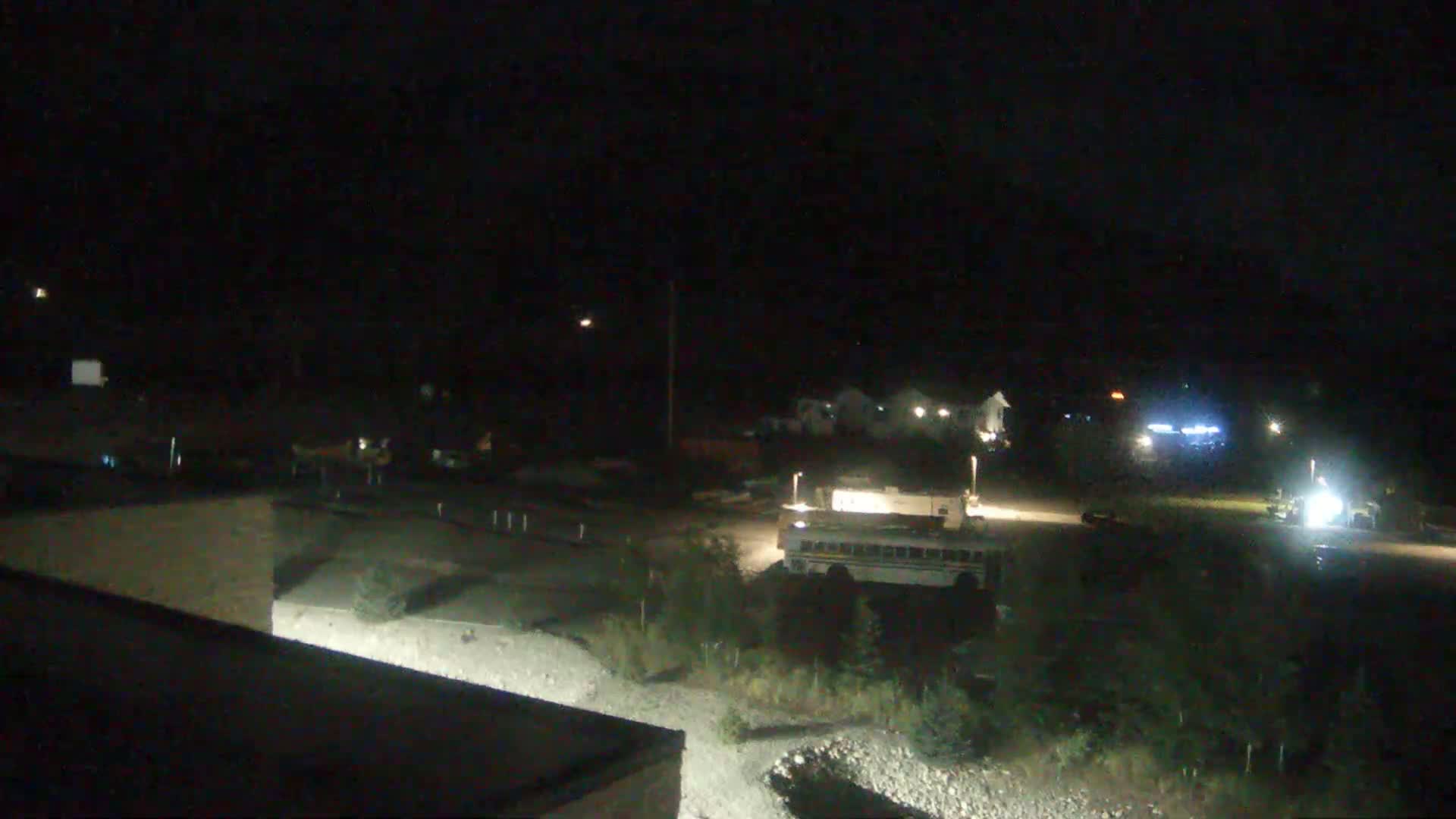 A nighttime view shows a school bus and other vehicles in a mostly empty parking lot near some buildings under a dark sky.