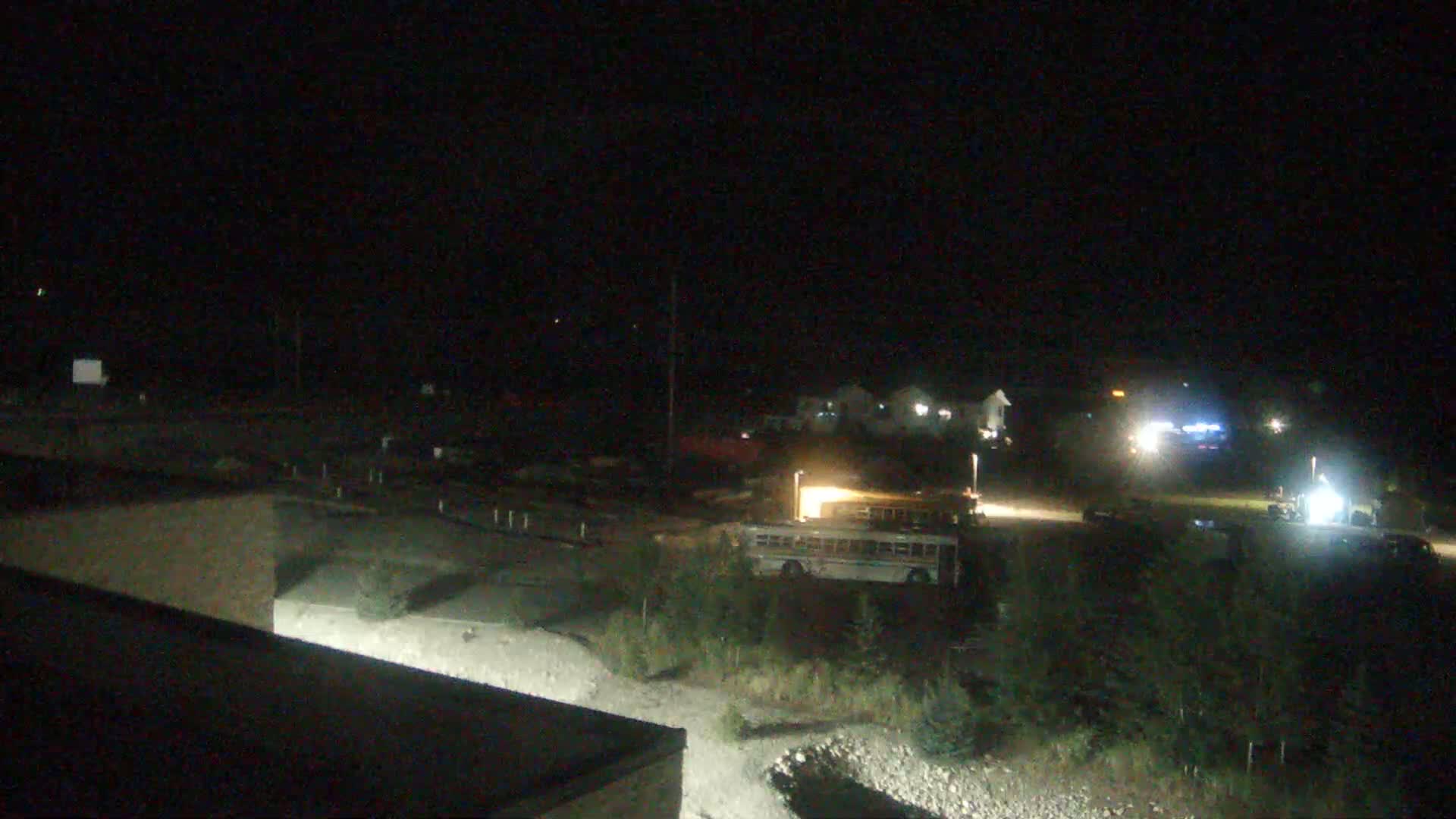 A nighttime view of a parking lot with a school bus and other vehicles, surrounded by sparse vegetation under a clear, dark sky.