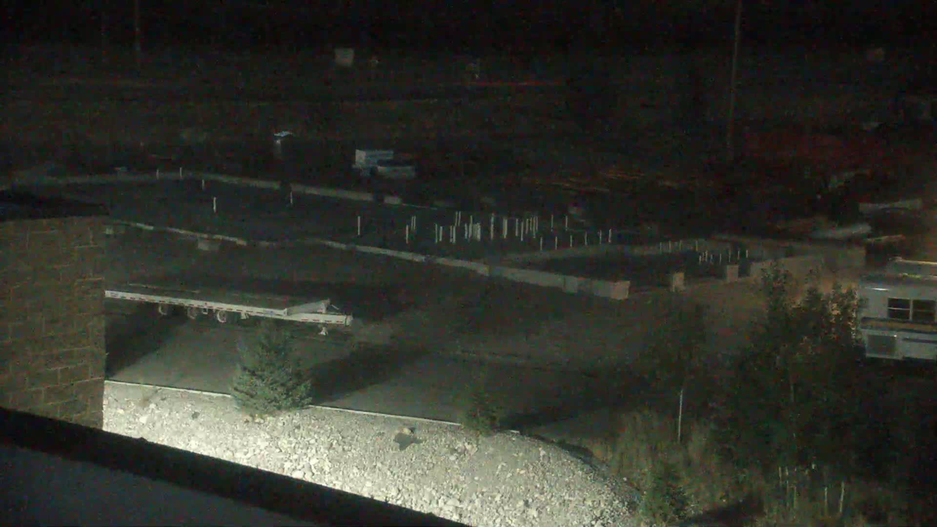 A nighttime, high-angle view shows a flatbed trailer parked near a partially-constructed building site, with a rocky area and some vegetation in the foreground.