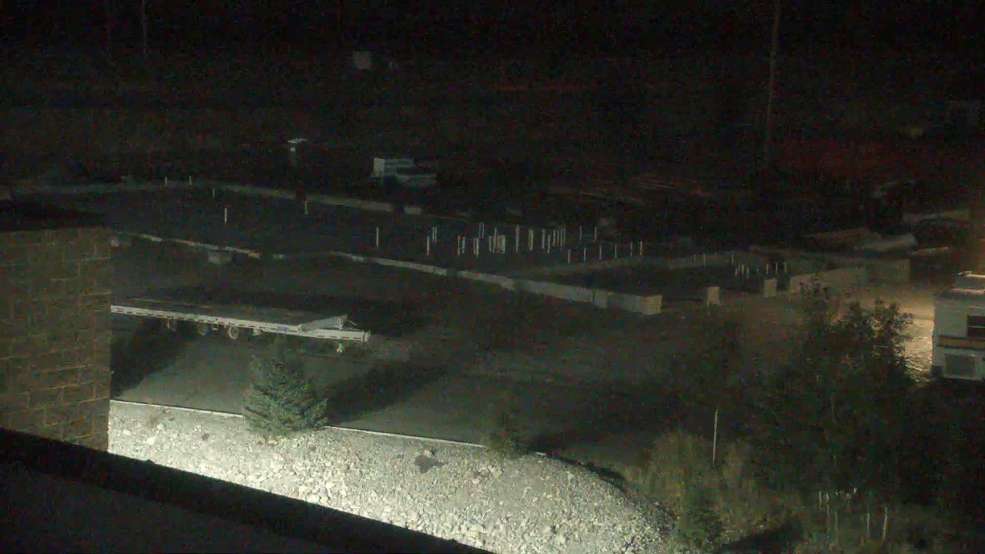 A nighttime, low-angle view shows a flat, paved area with concrete foundations, a small evergreen tree, and a portion of a large trailer, all under dark conditions.