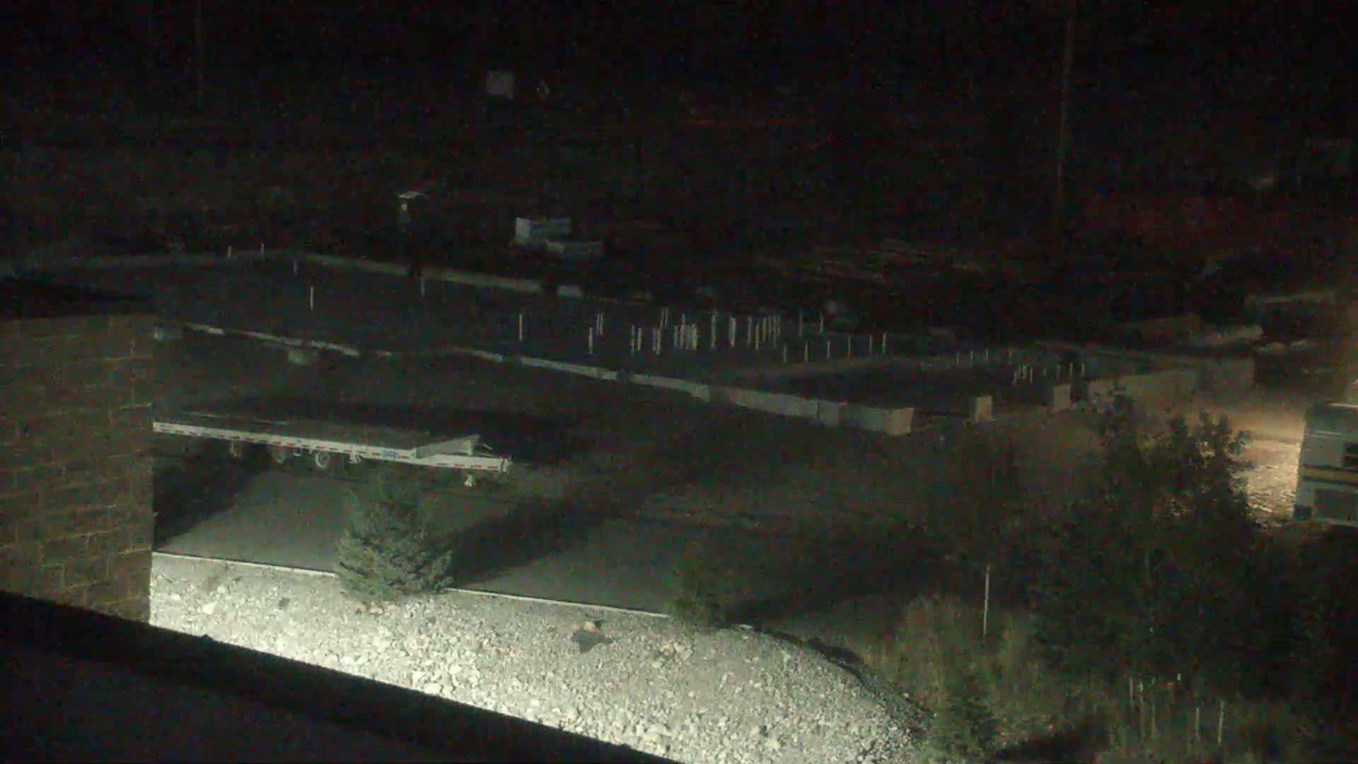 A nighttime, low-light view shows a flatbed trailer parked near a construction site with numerous posts and what appears to be a gravel area.