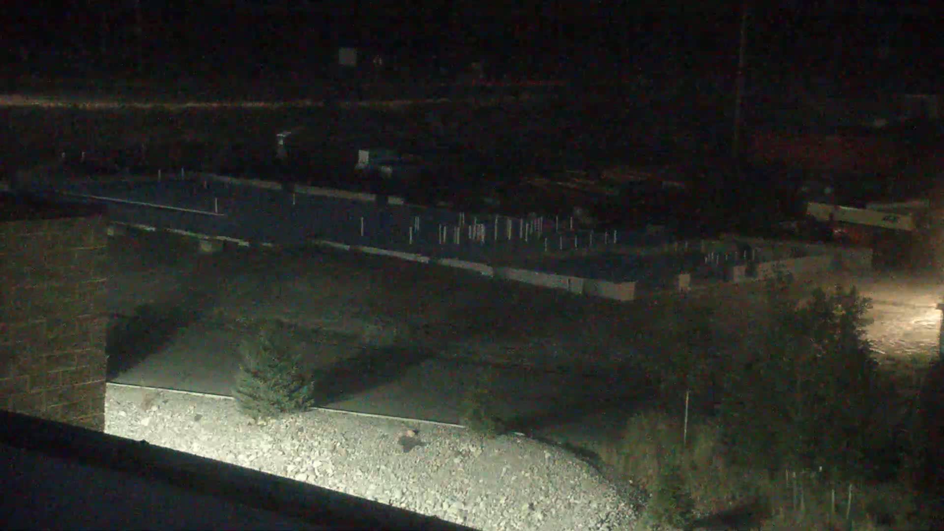 A nighttime, low-light view shows a flatbed trailer parked near a construction site with numerous posts and what appears to be a gravel area.