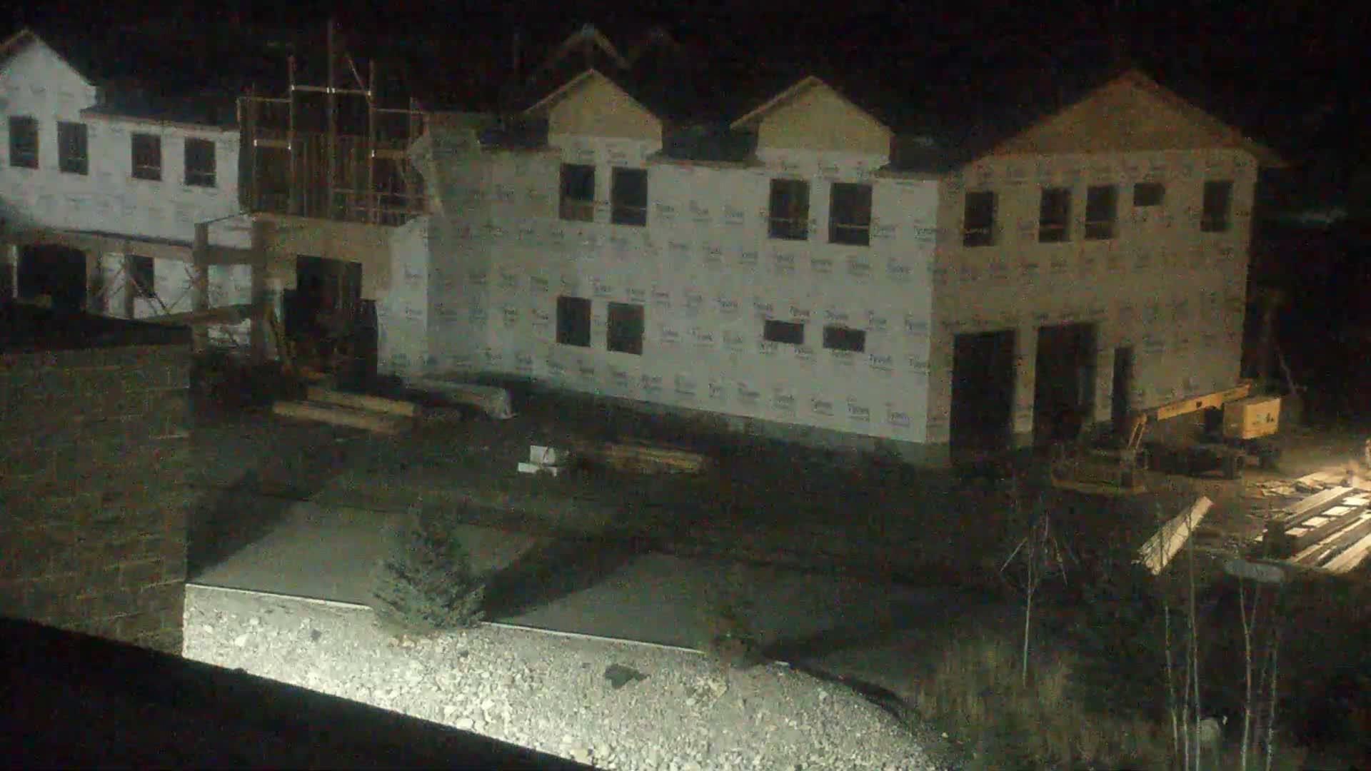 At night, several multi-story houses under construction, wrapped in white weather barrier and featuring some exposed framing, are visible alongside construction equipment and materials on the ground under clear skies.