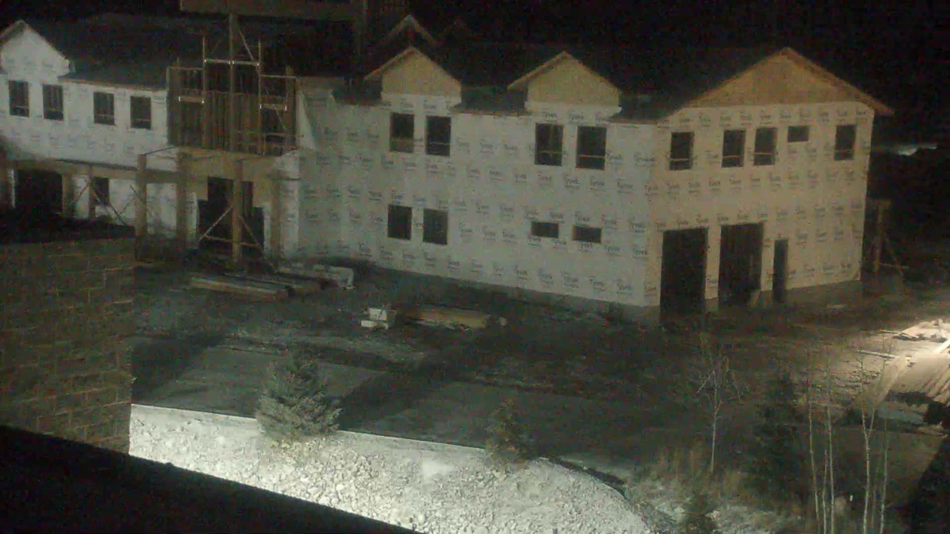 A large residential building under construction, covered in white house wrap with several roof gables and framed openings for windows and doors, is seen at night under clear skies, with construction debris and sparse vegetation on the dark, uneven ground.