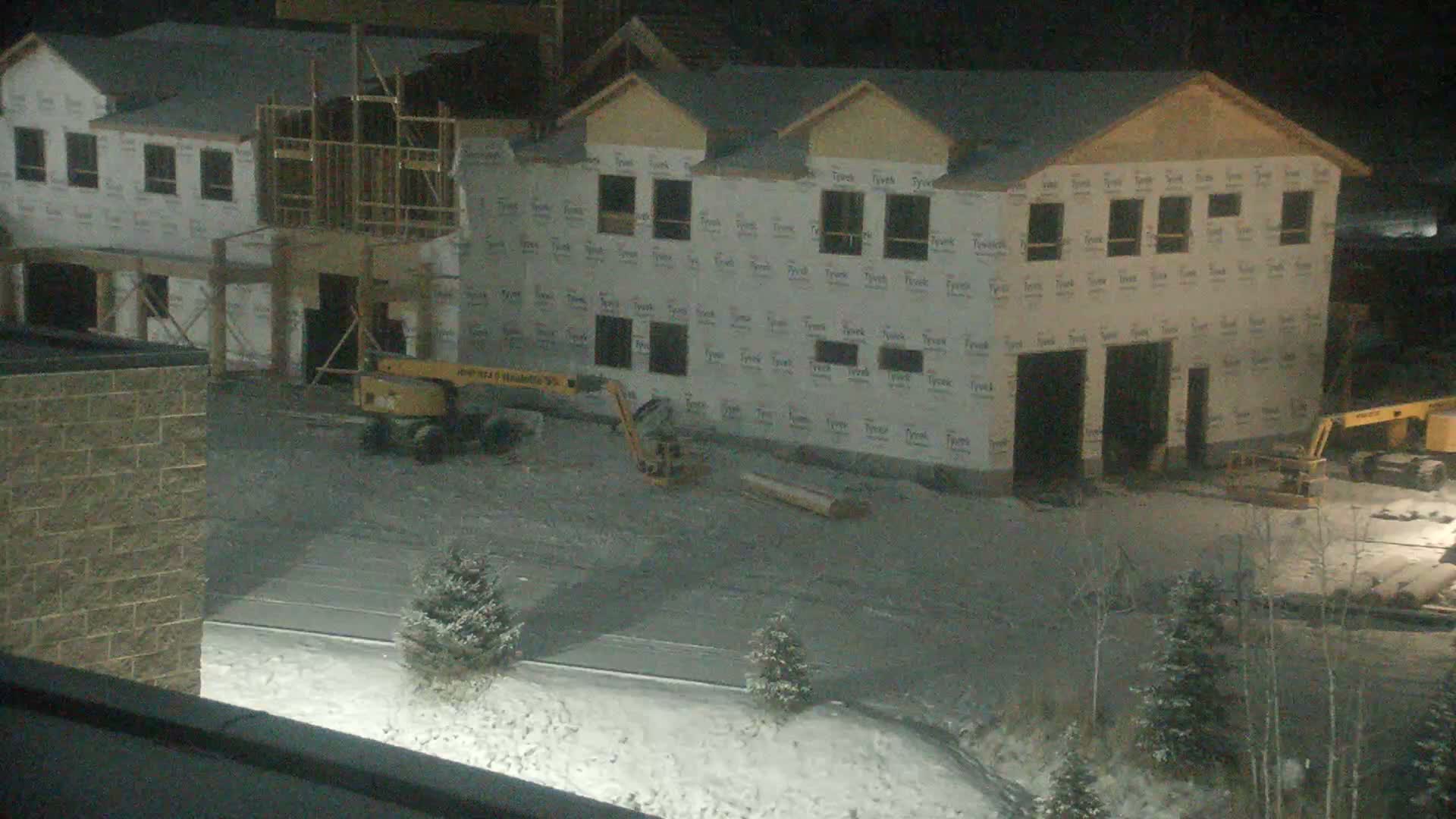 A dark, aerial view shows a construction site at night with several multi-story buildings in various stages of framing and sheathing, two yellow construction vehicles parked on unpaved ground, and a partially snow-covered landscape.
