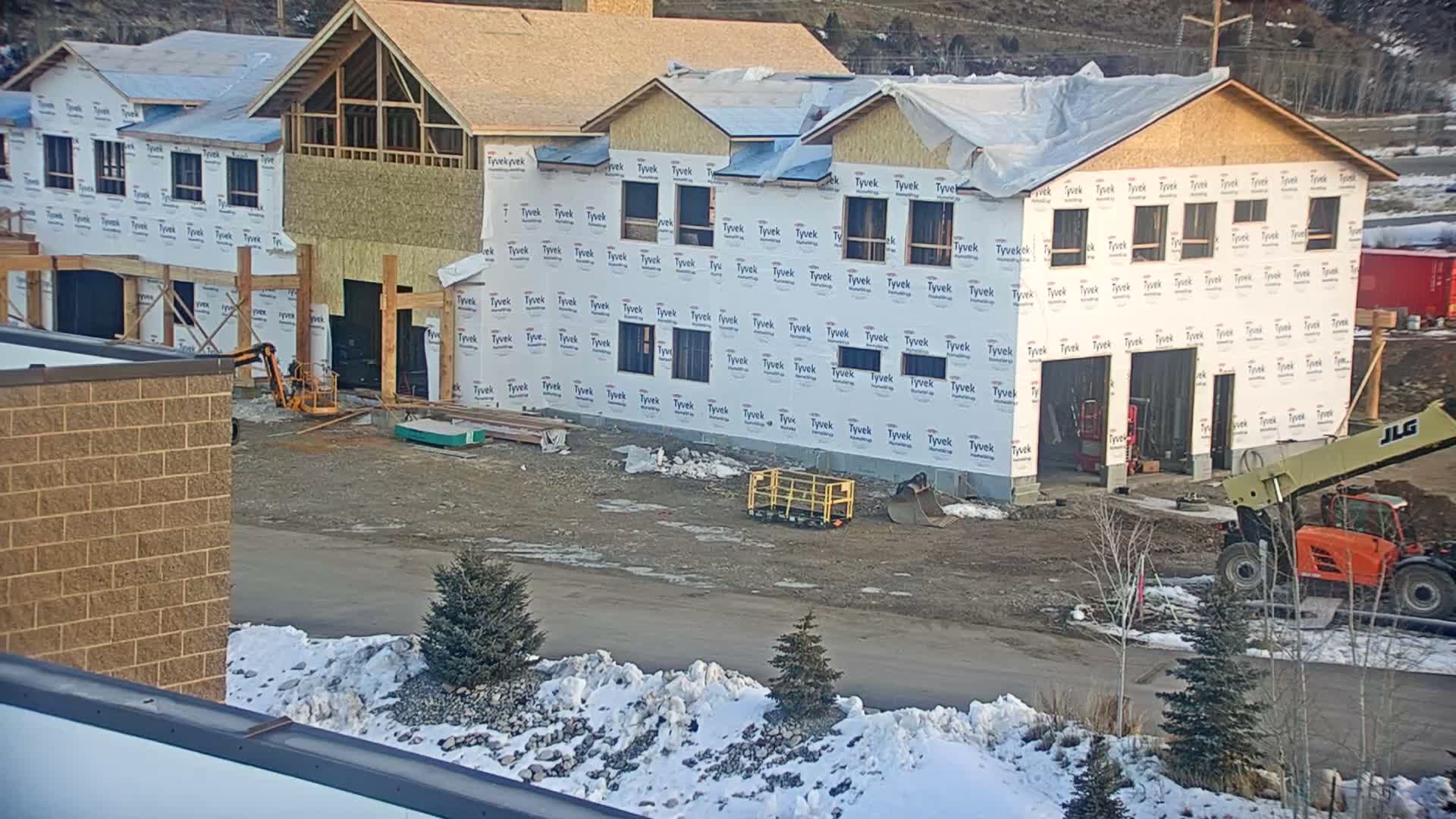 A dark, aerial view shows a construction site at night with several multi-story buildings in various stages of framing and sheathing, two yellow construction vehicles parked on unpaved ground, and a partially snow-covered landscape.