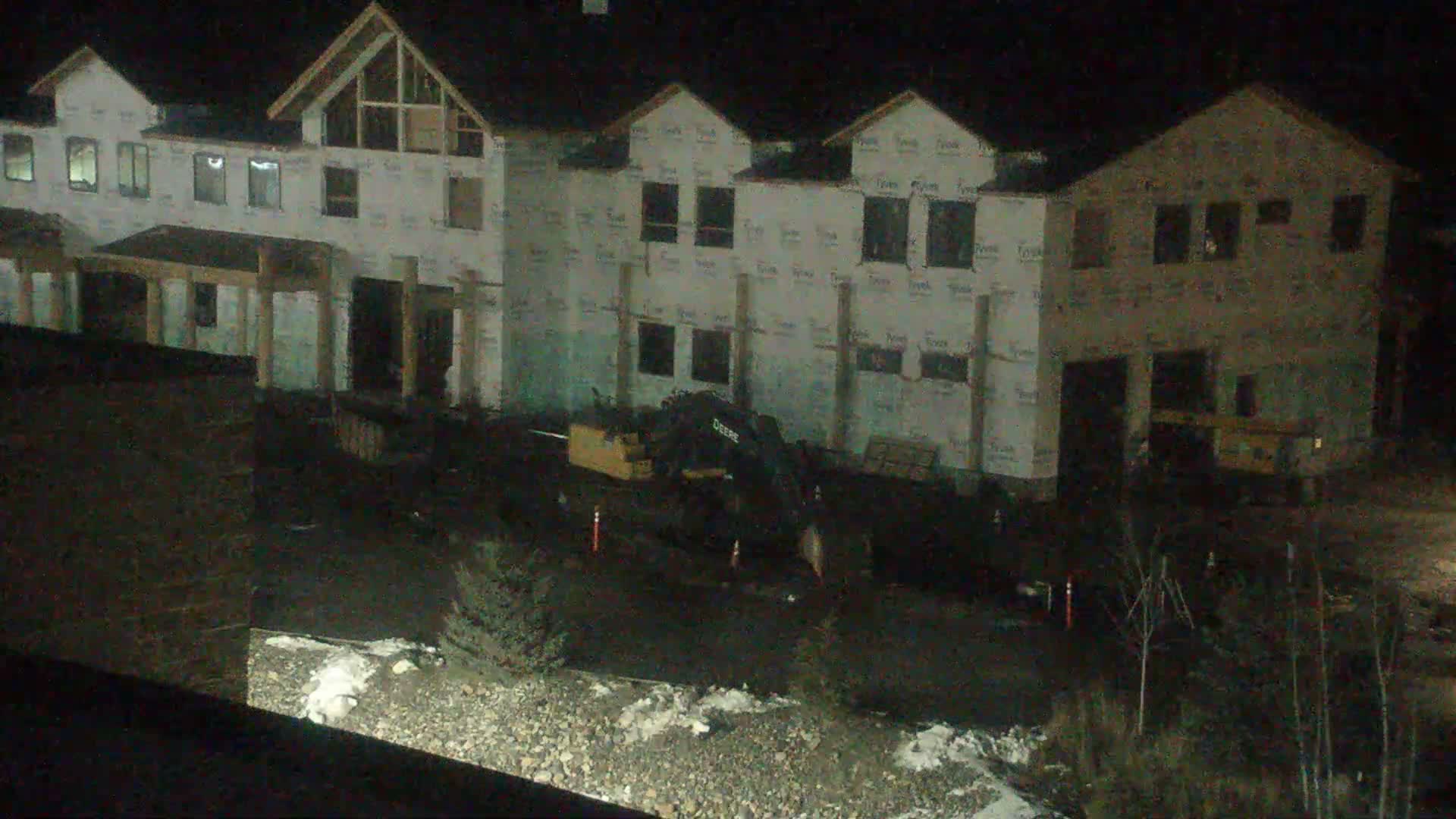 At night, a construction site is visible with multiple multi-story buildings in various stages of framing and sheathing, an excavator on the muddy ground, and patches of snow under clear skies.