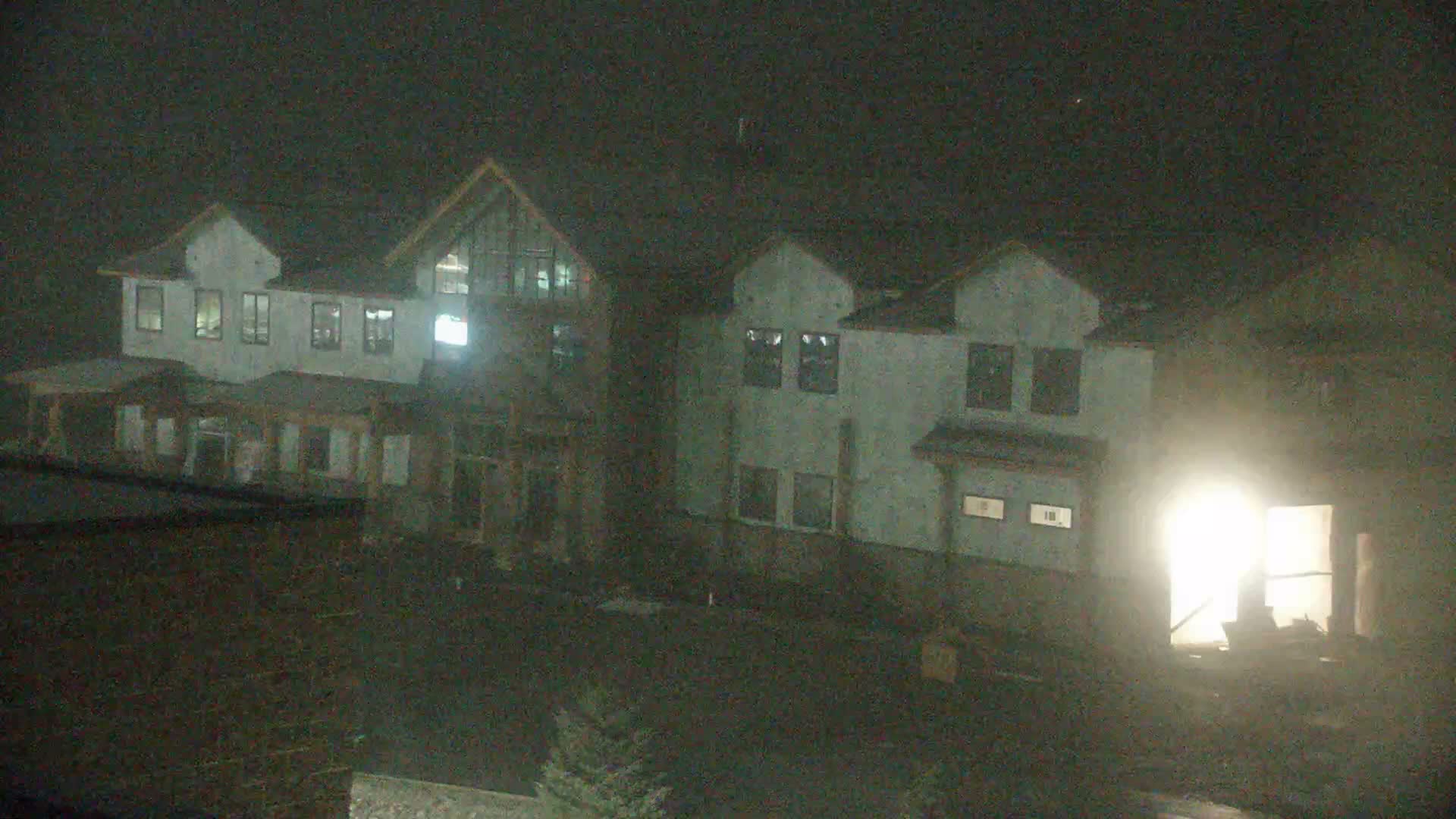 At night, a construction site is visible with multiple multi-story buildings in various stages of framing and sheathing, an excavator on the muddy ground, and patches of snow under clear skies.