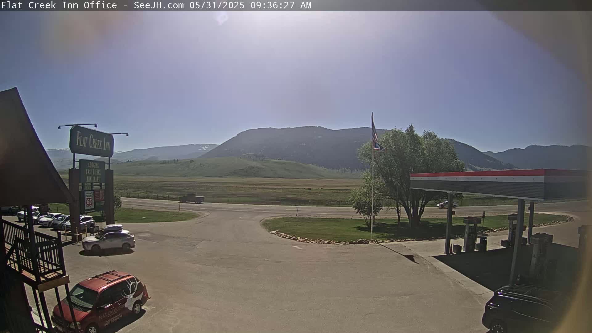 A sunny day reveals a parking lot with several vehicles near a gas station and inn, set against a backdrop of mountains and a flat, grassy plain.