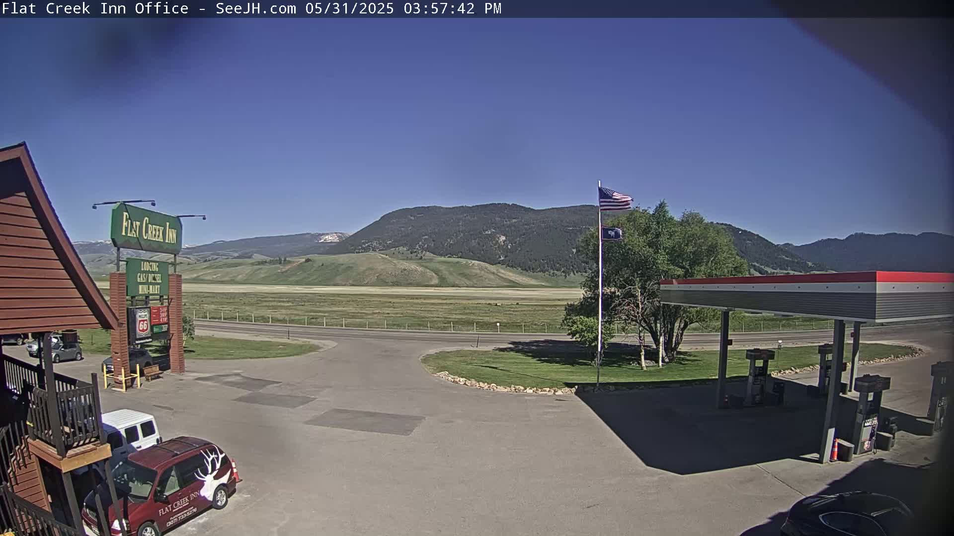 A gas station and inn parking lot are visible, under a clear blue sky, with a mountain range in the background.