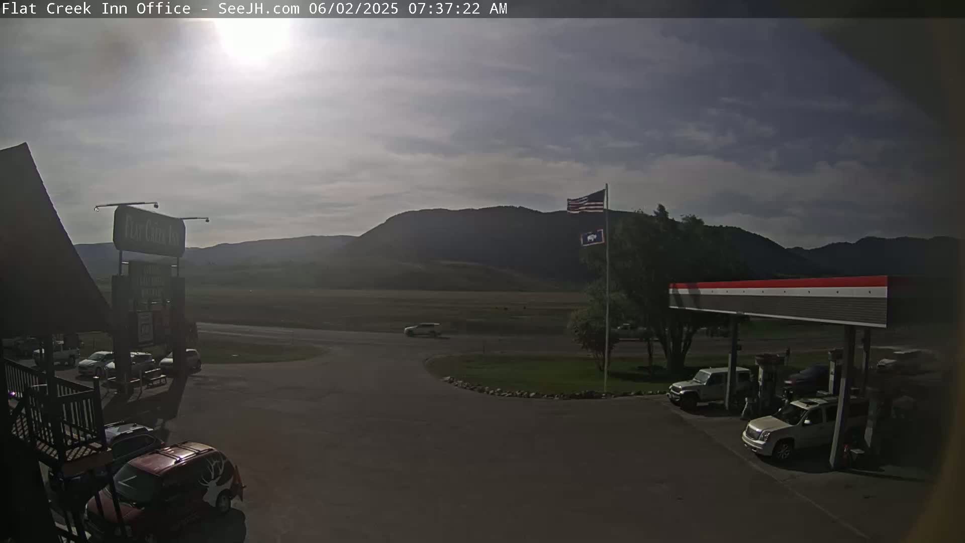 A mostly cloudy morning scene shows a gas station and parking lot next to a building, with a few vehicles visible, in front of flat land and distant mountains.