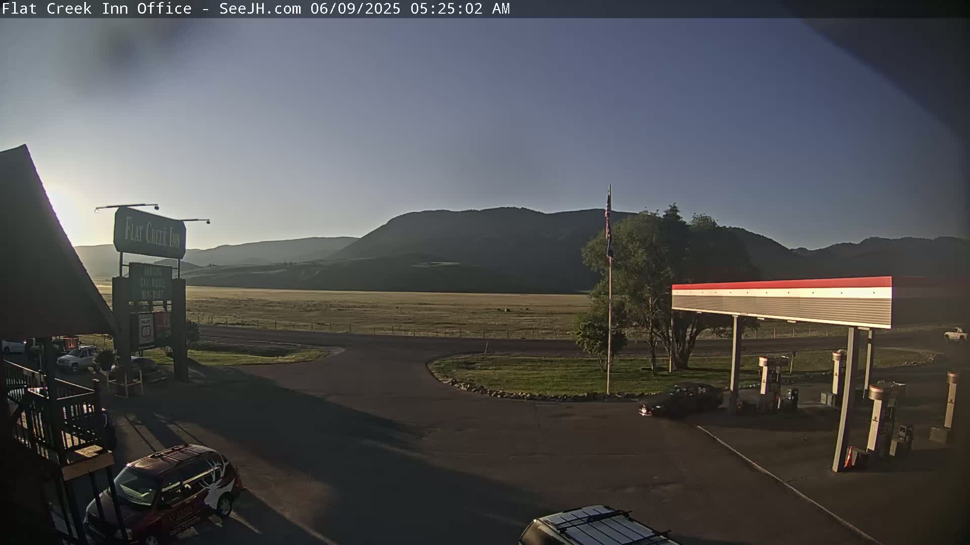 A gas station and motel are situated in a flat, grassy area before a range of mountains under a clear, early morning sky.