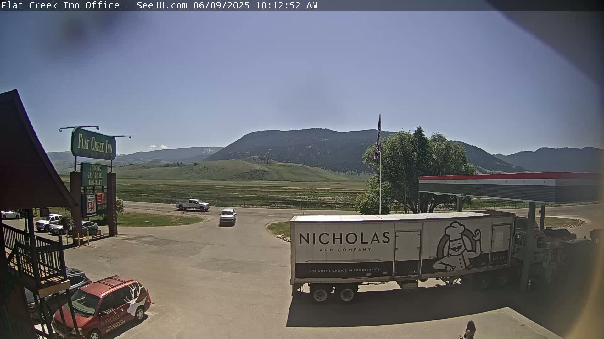 A sunny day shows a parking lot with several vehicles, a large food service truck, a gas station canopy, and a Flat Creek Inn sign in the foreground, with mountains visible in the background.