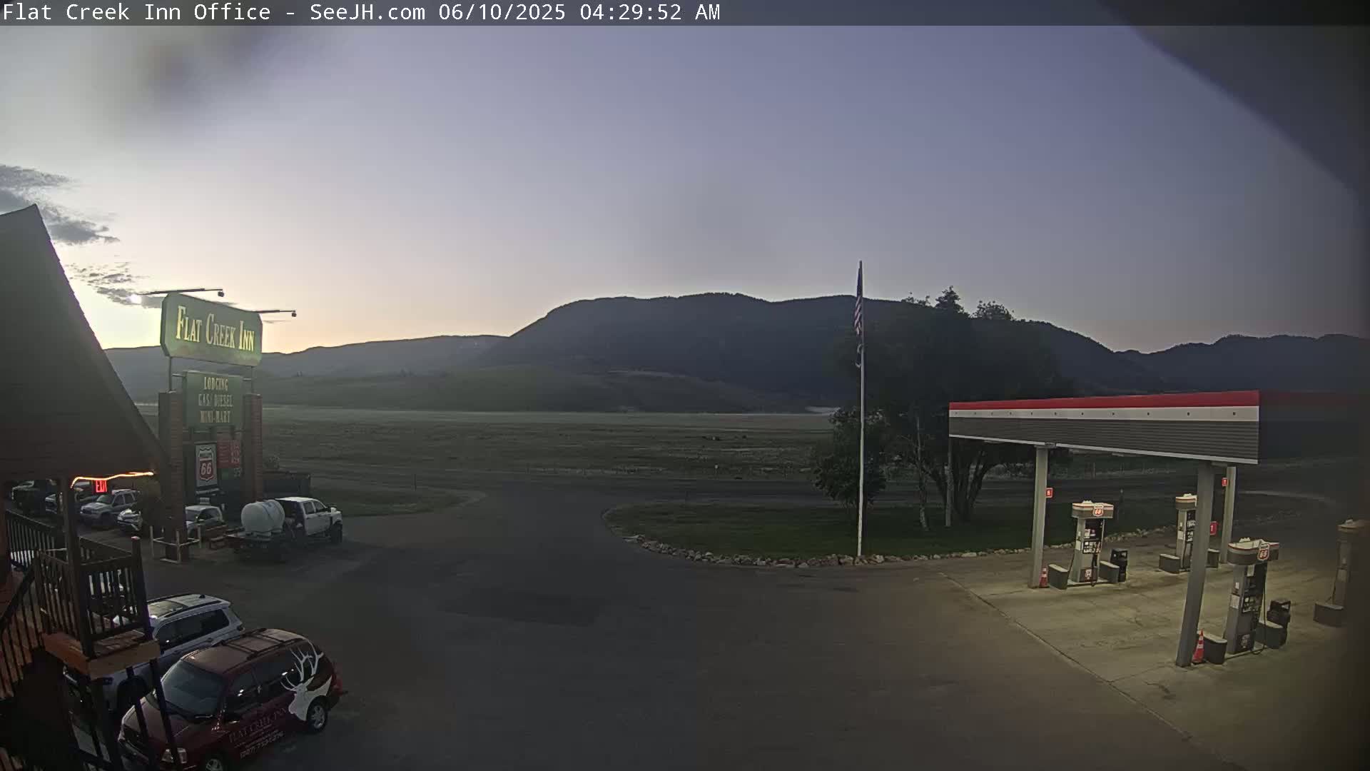 A gas station and motel are situated in a flat, grassy area at dawn, with mountains visible in the background under a clear, dusky sky.