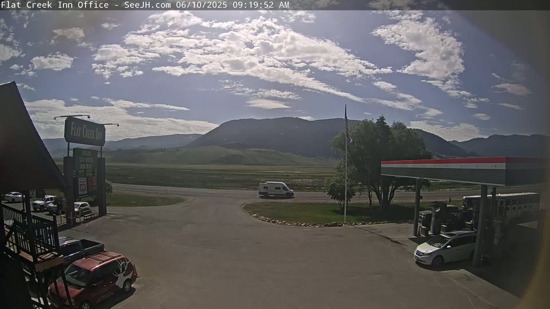 A mostly sunny day with some clouds over a valley, showing a gas station, inn, and several vehicles parked near a road.