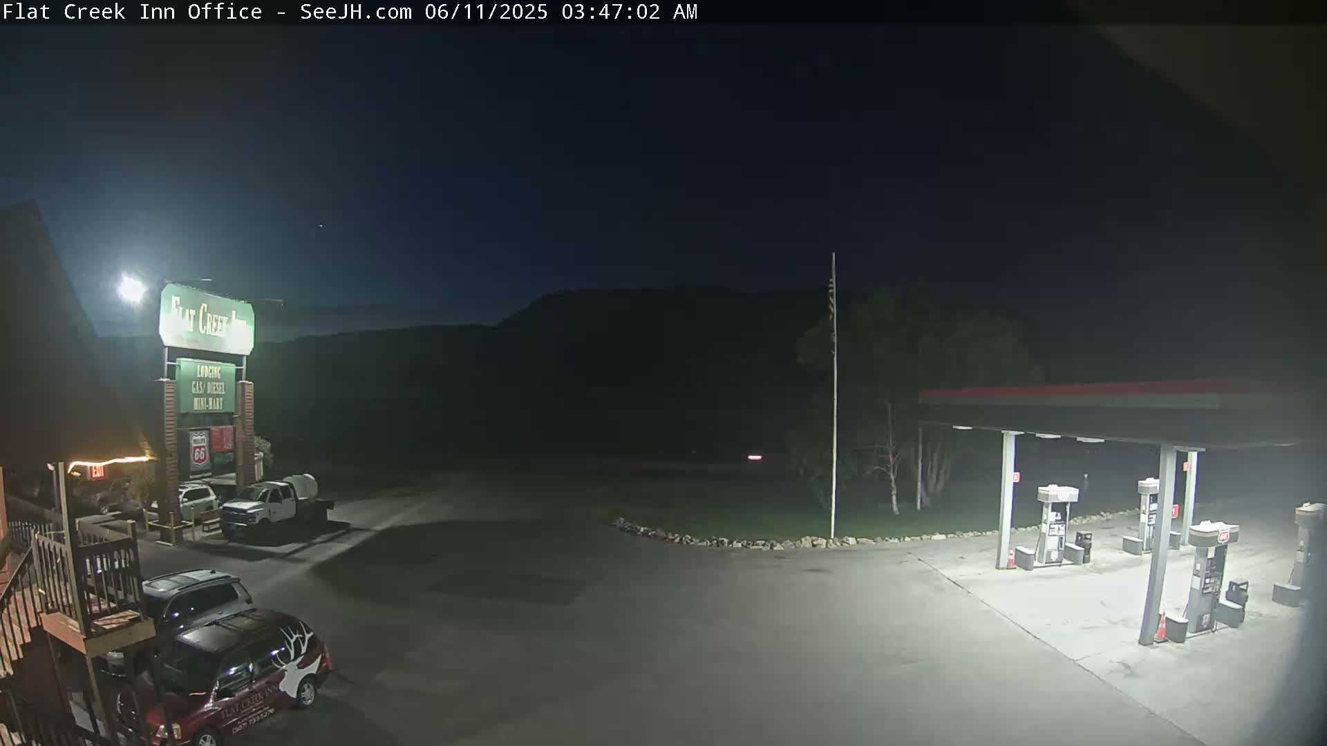 A nighttime view of a gas station and motel parking lot with several vehicles parked, under a dark, clear sky.