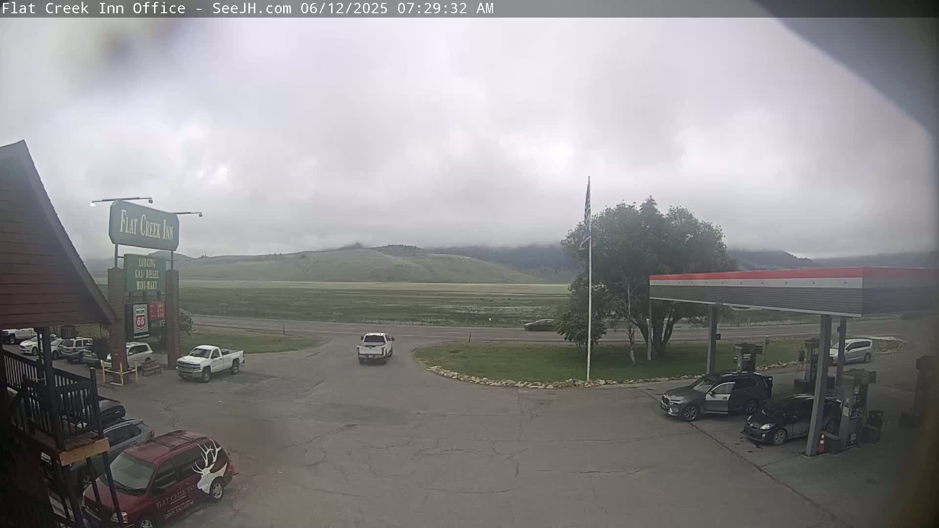 A mostly cloudy, overcast day shows a gas station and inn parking lot with several vehicles, and a view of a flat, green valley with distant mountains.