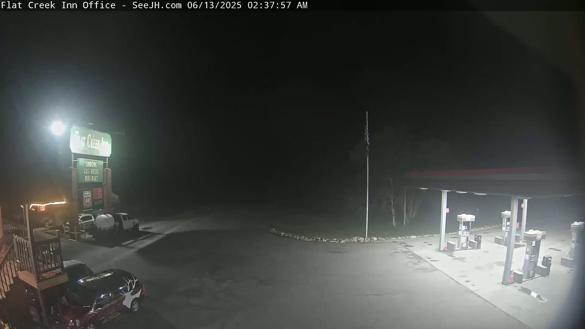 A nighttime view of a gas station and motel parking lot with several vehicles parked, under dark, clear skies.