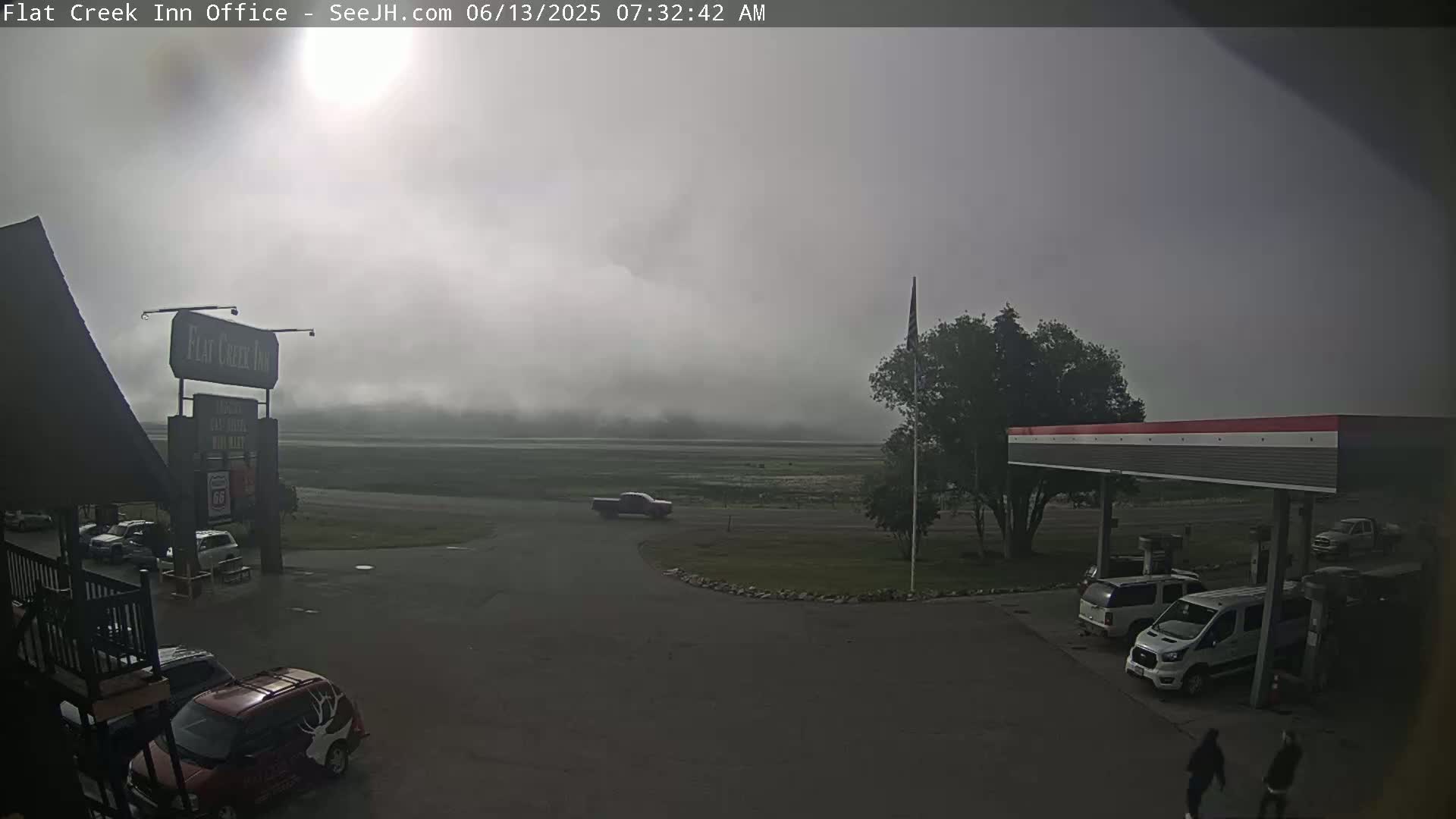 A foggy morning scene shows a parking lot with several vehicles near a gas station and inn, with a flat, low-lying landscape visible in the distance.