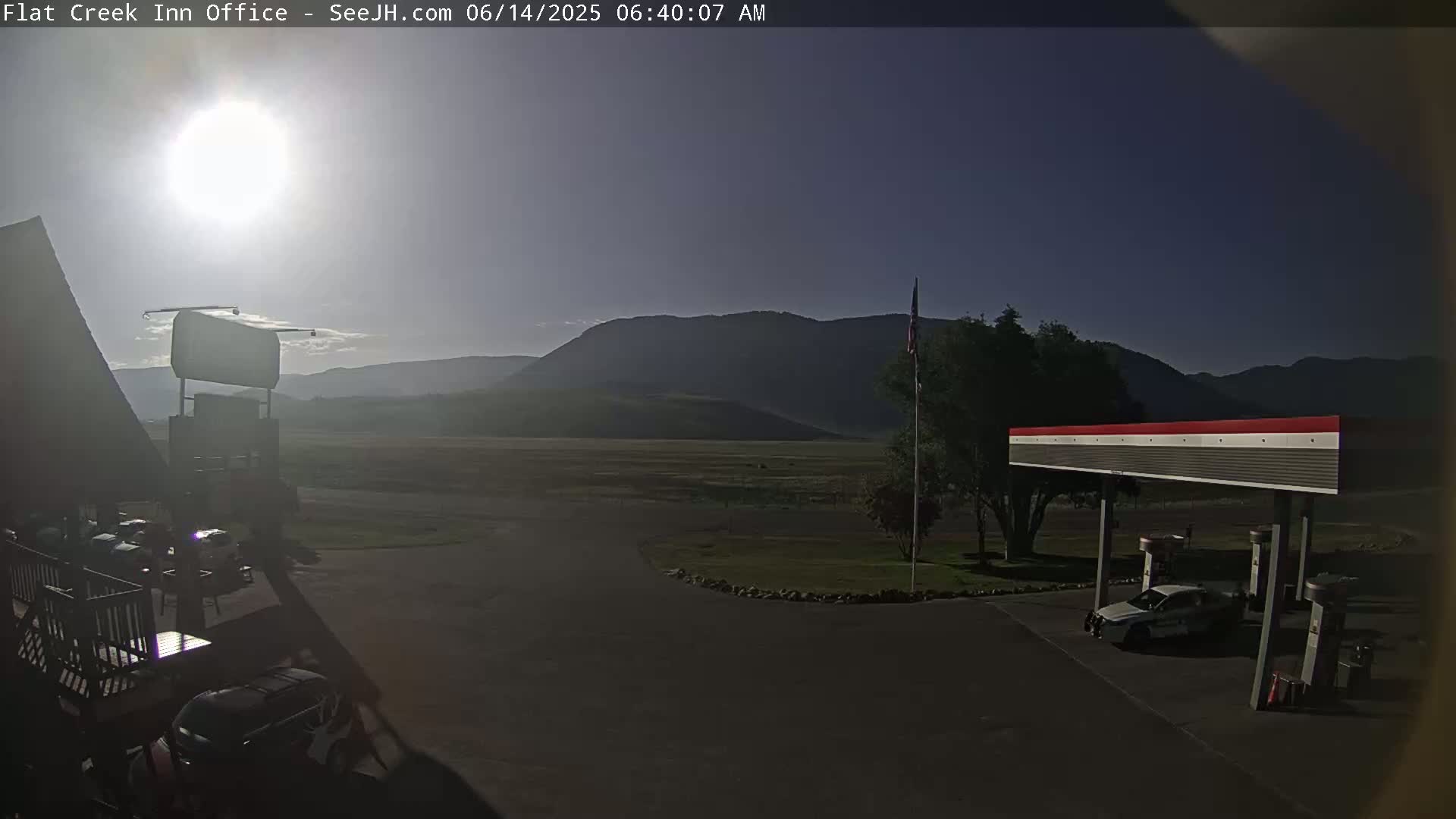A sunny morning view shows a gas station and several vehicles parked in a lot, with mountains visible in the background.
