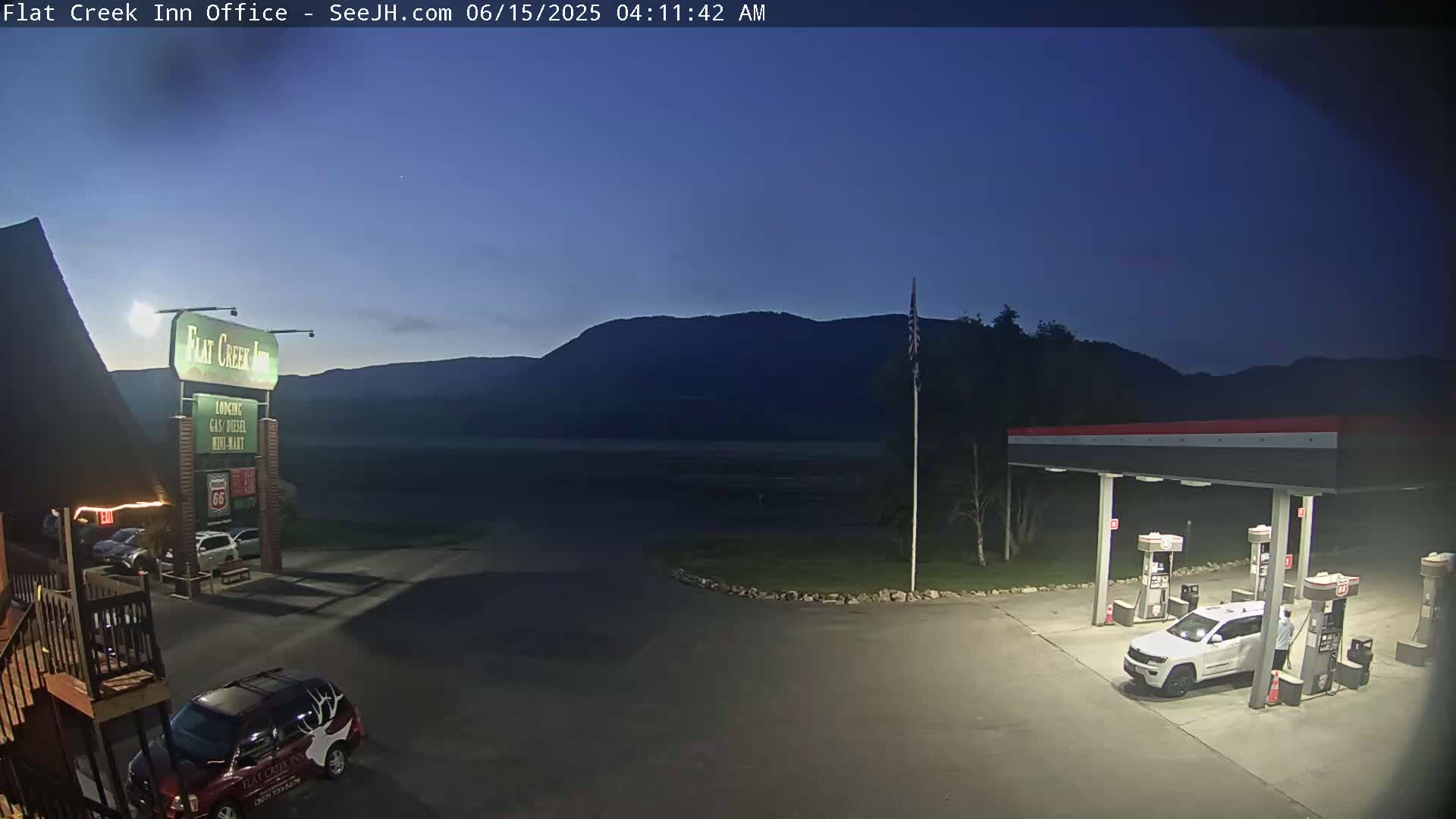 A gas station and motel are illuminated at night under a dark blue, pre-dawn sky, with mountains visible in the background.
