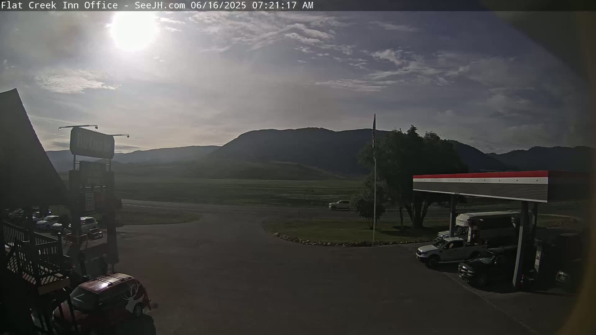 A sunny, partly cloudy day reveals a parking lot with several vehicles near a gas station, set against a backdrop of distant, dark mountains and a flat, green field.