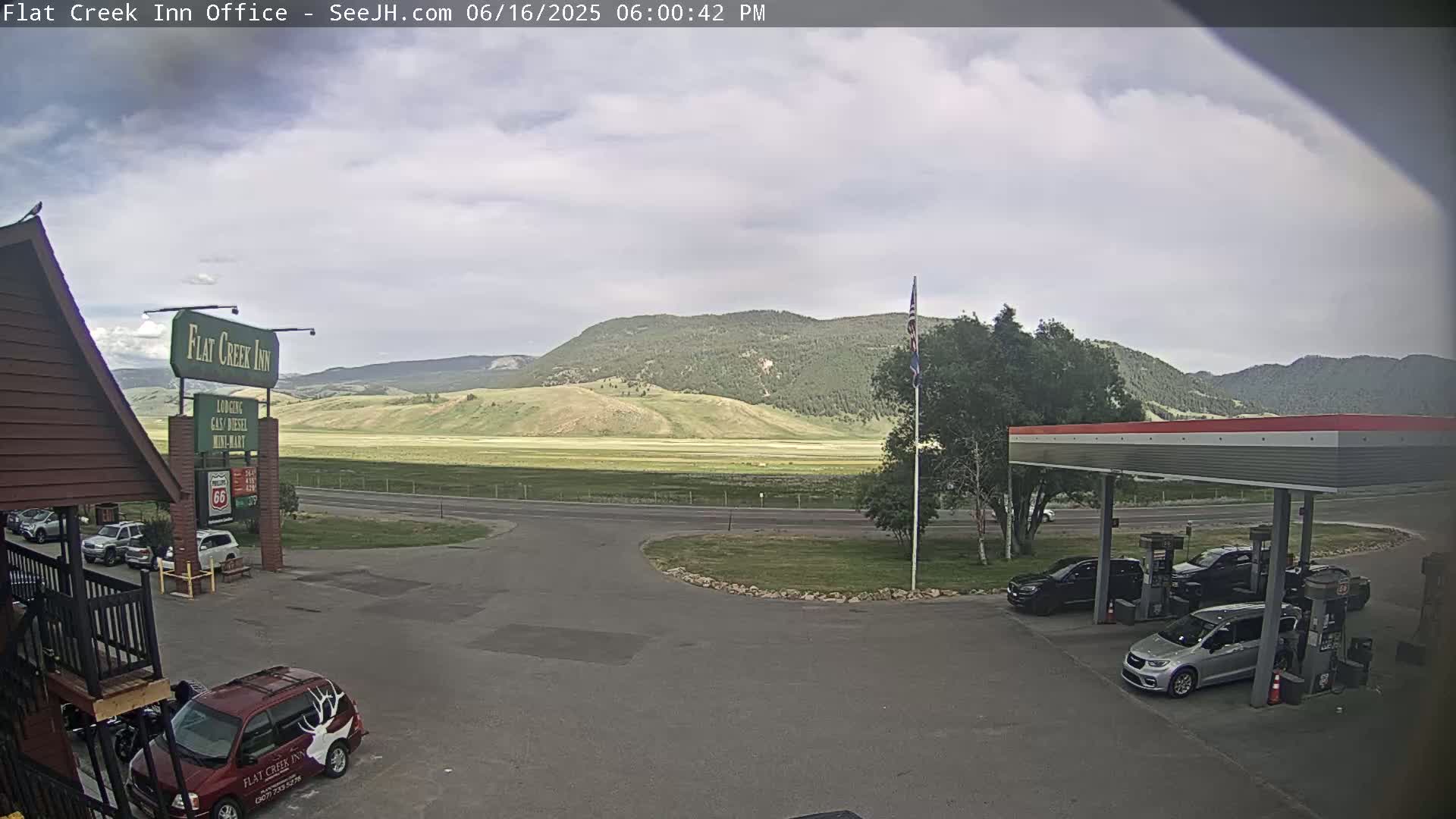 A gas station and motel parking lot are situated before a broad, grassy valley and distant mountains under a partly cloudy sky.