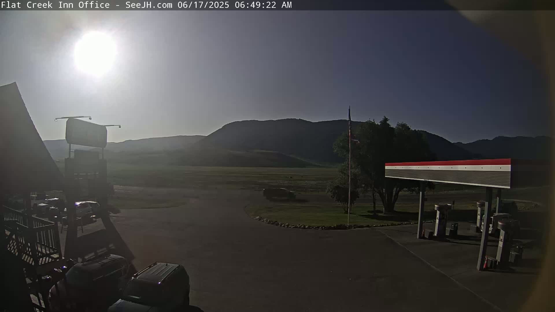 A sunny morning scene shows a gas station and a parking lot with several vehicles, situated before a flat expanse of land and distant mountains.