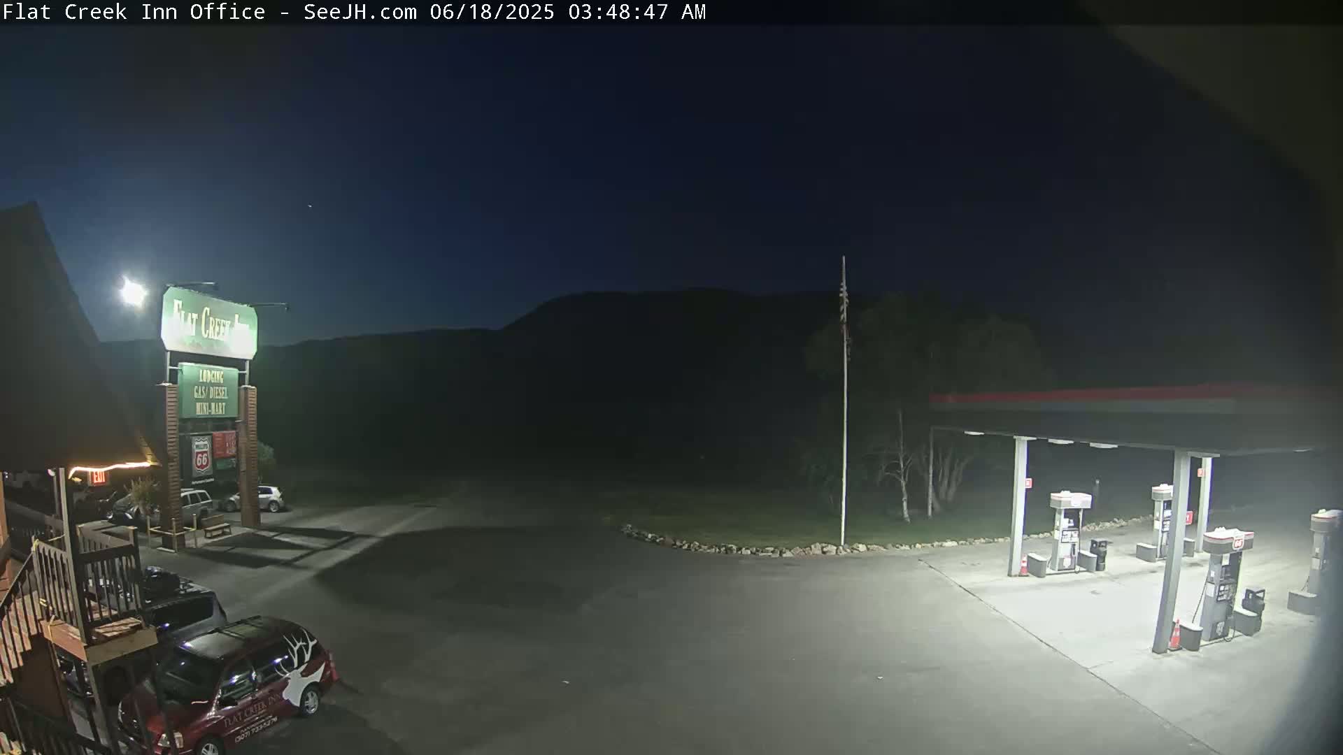 A nighttime view of a gas station and motel parking lot, illuminated by artificial lights, with dark mountains in the background under a clear, dark sky.